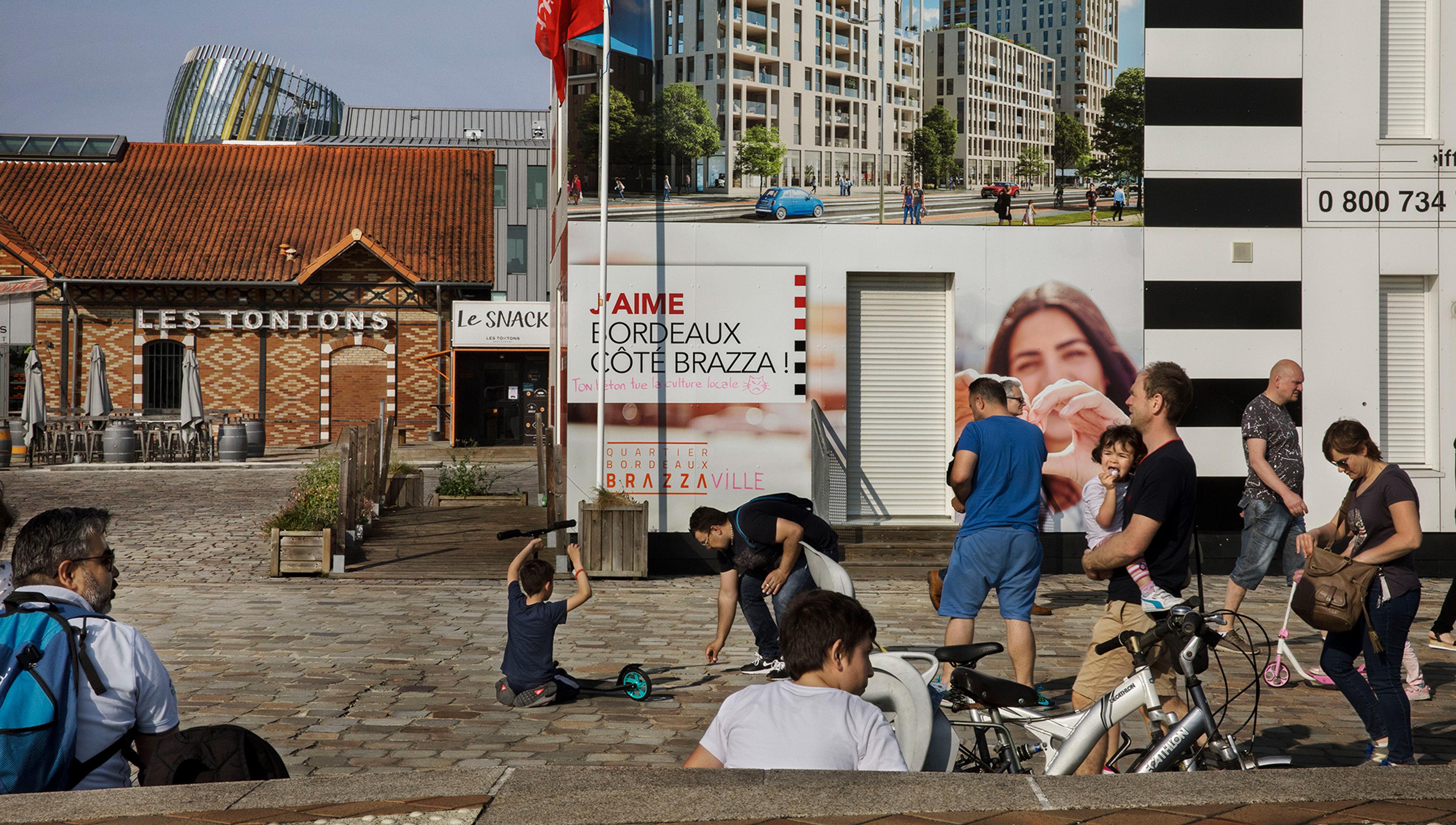 People sitting and walking through a city square on a sunny day, with a red brick building and large advertising posters covering other buildings.