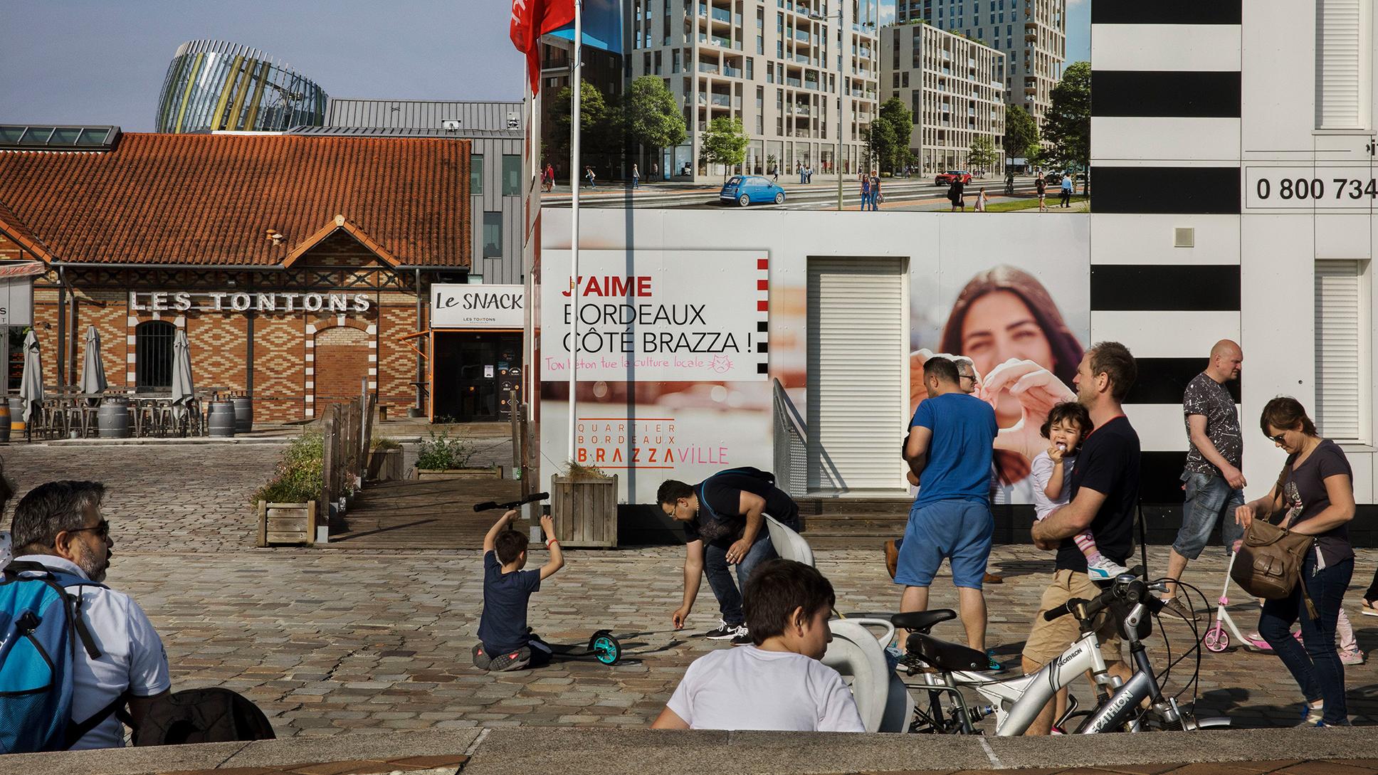 People sitting and walking through a city square on a sunny day, with a red brick building and large advertising posters covering other buildings.