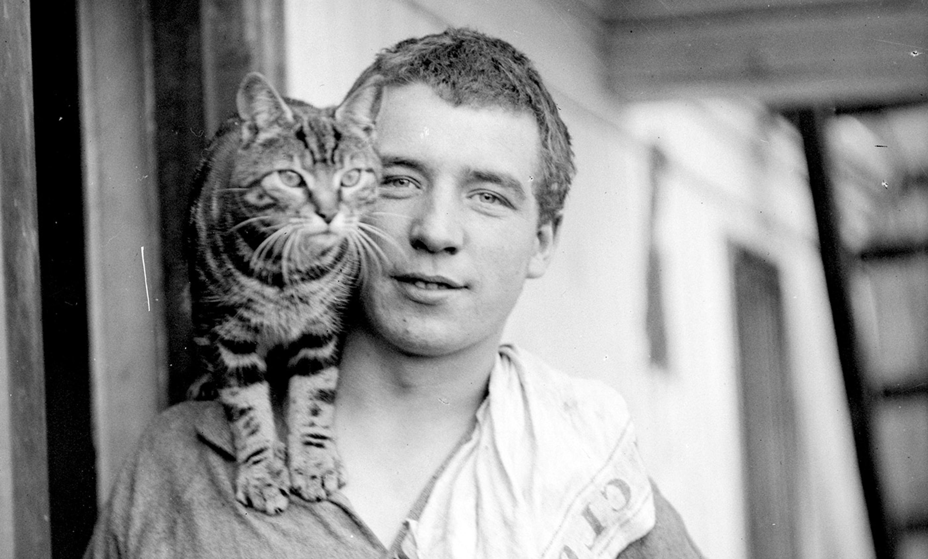Black and white photo of a man with a tabby cat perched on his shoulder, standing in front of a building’s doorway.
