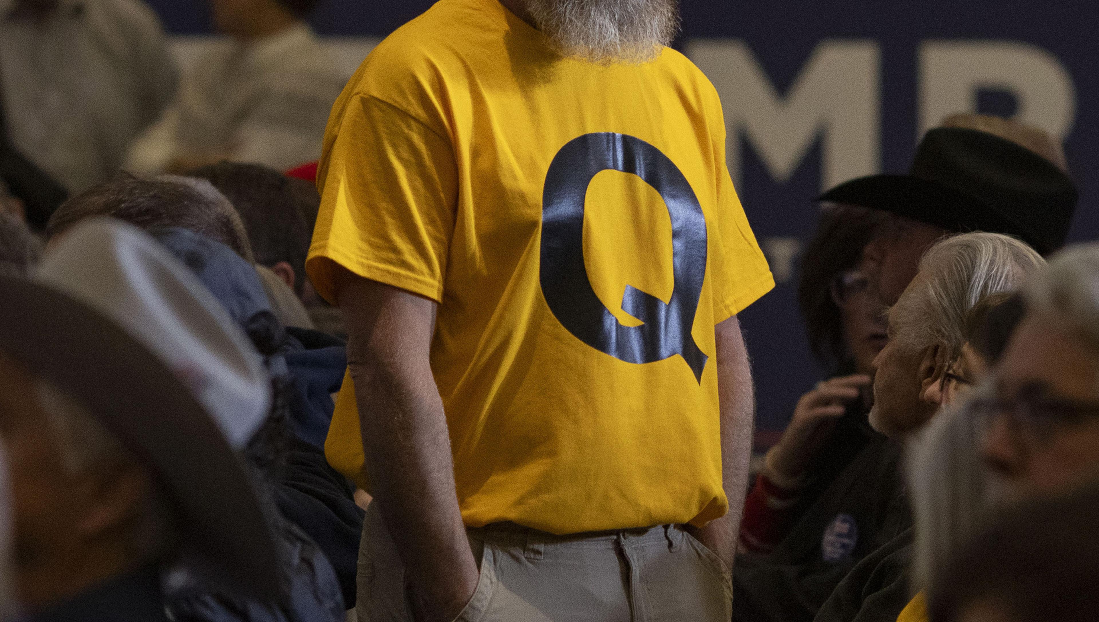 Photo of a man in a yellow T-shirt with a large black letter “Q” standing among seated people at an indoor event.