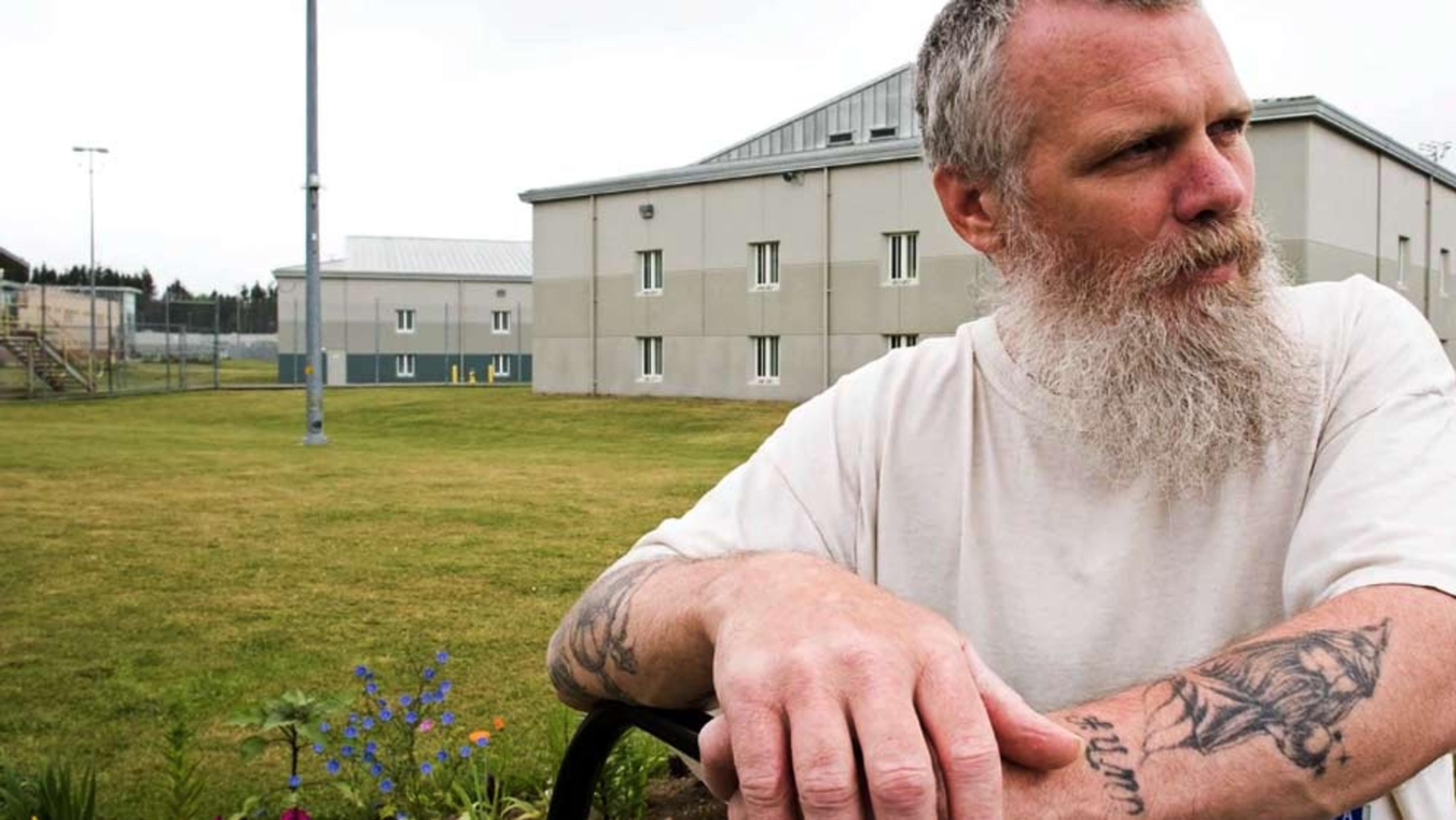 A man with a long beard and tattoos leaning on a railing outside a grey building in a fenced area with a grassy field.
