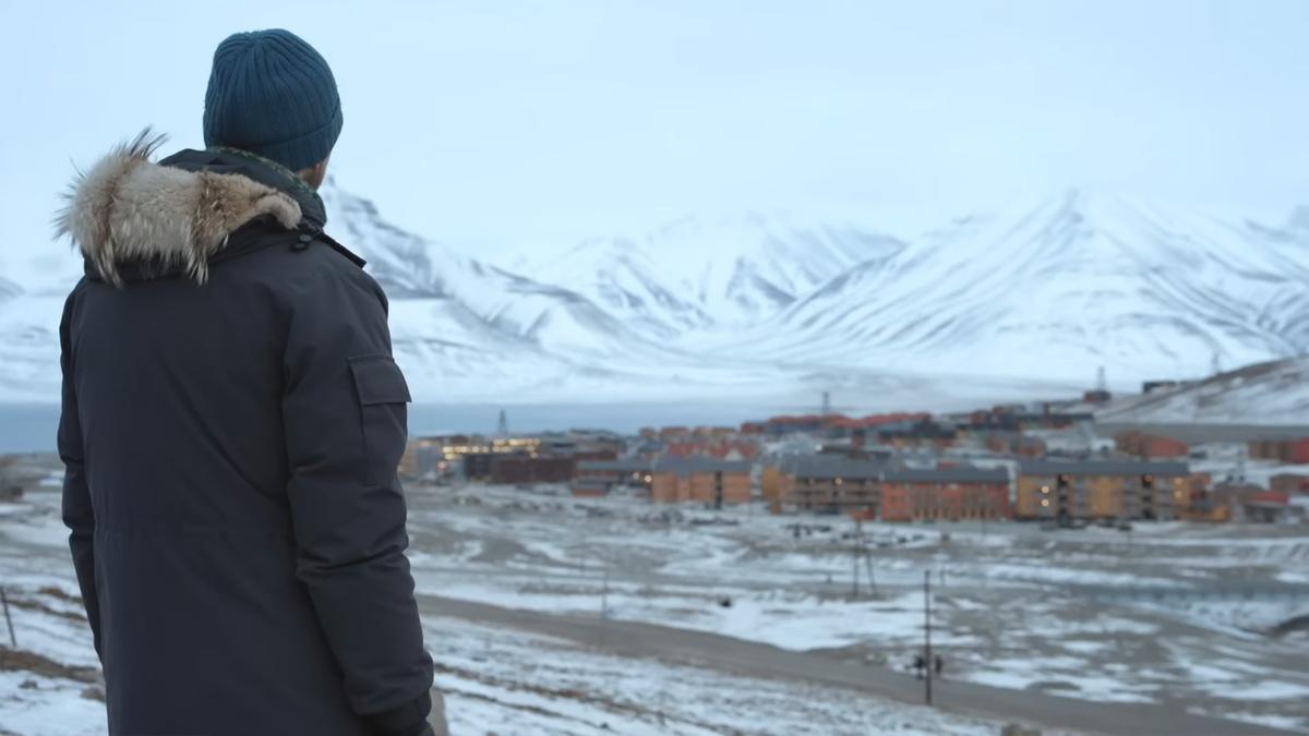 Photo of a person in a winter coat and beanie looking at a snow-covered town, with mountains in the background.