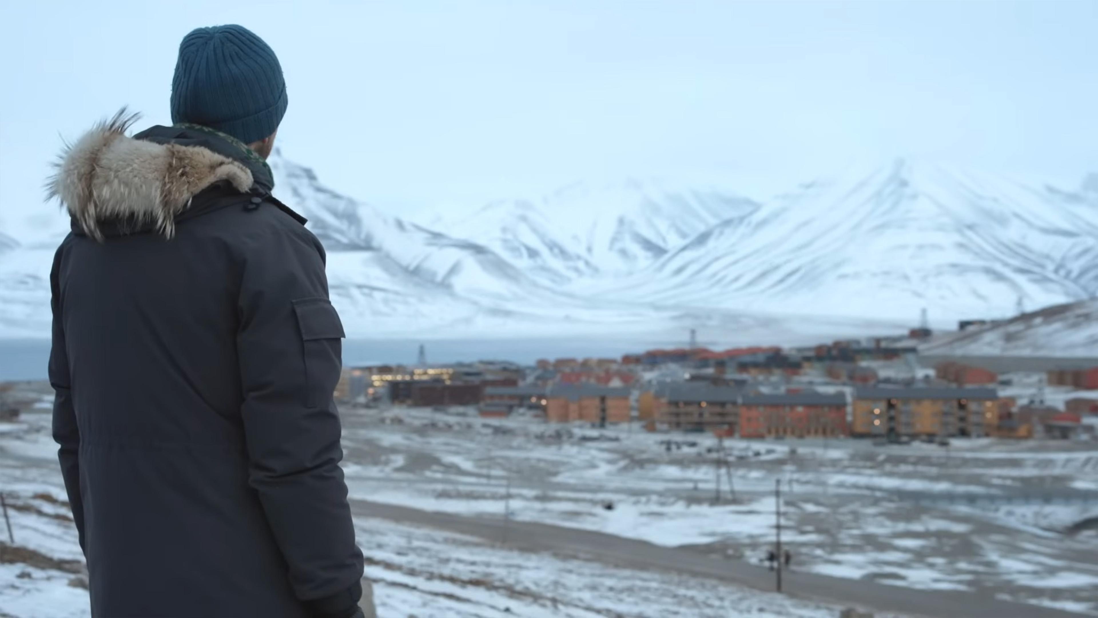 Photo of a person in a winter coat and beanie looking at a snow-covered town, with mountains in the background.