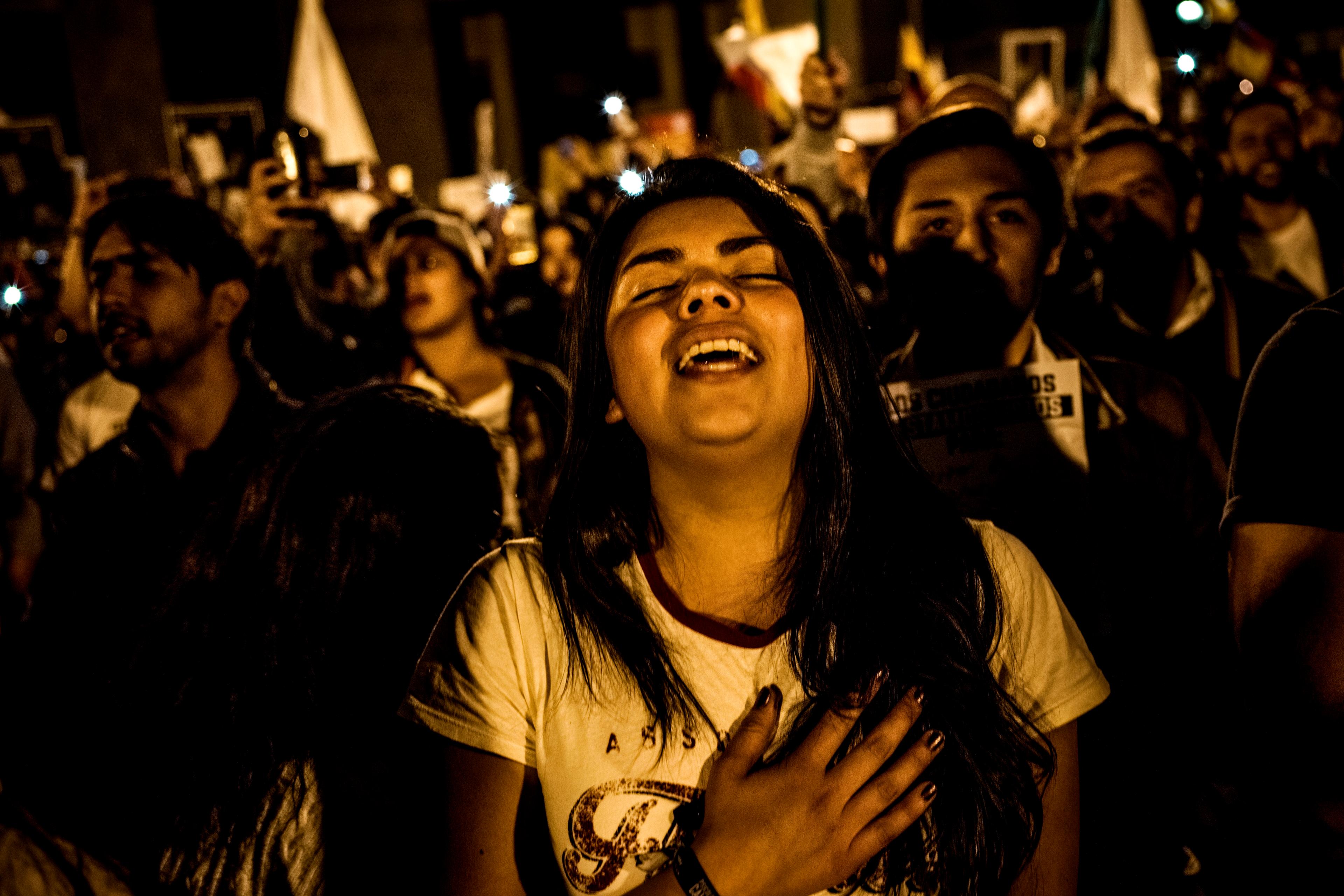 Photo of a crowd at night, a woman in the front smiles with her hand on her chest, others hold lights in the background.