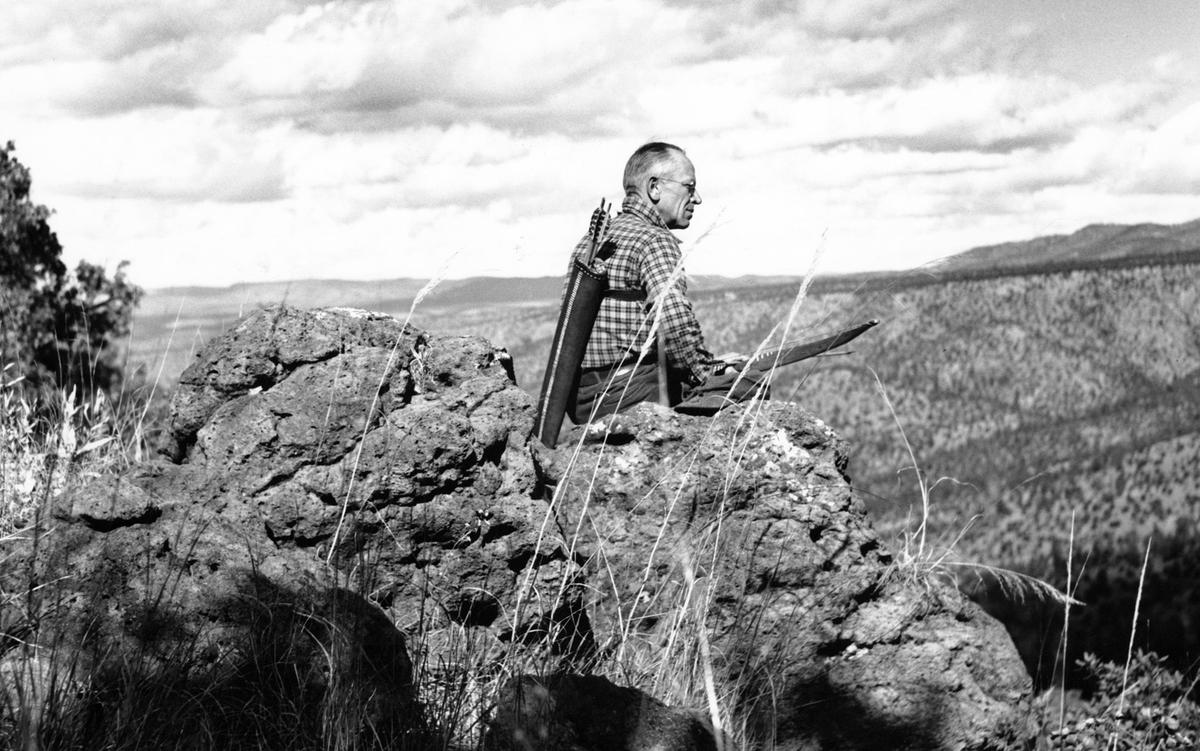 Black and white photo of a man with a bow and arrows sitting on a rocky outcrop overlooking a vast landscape.