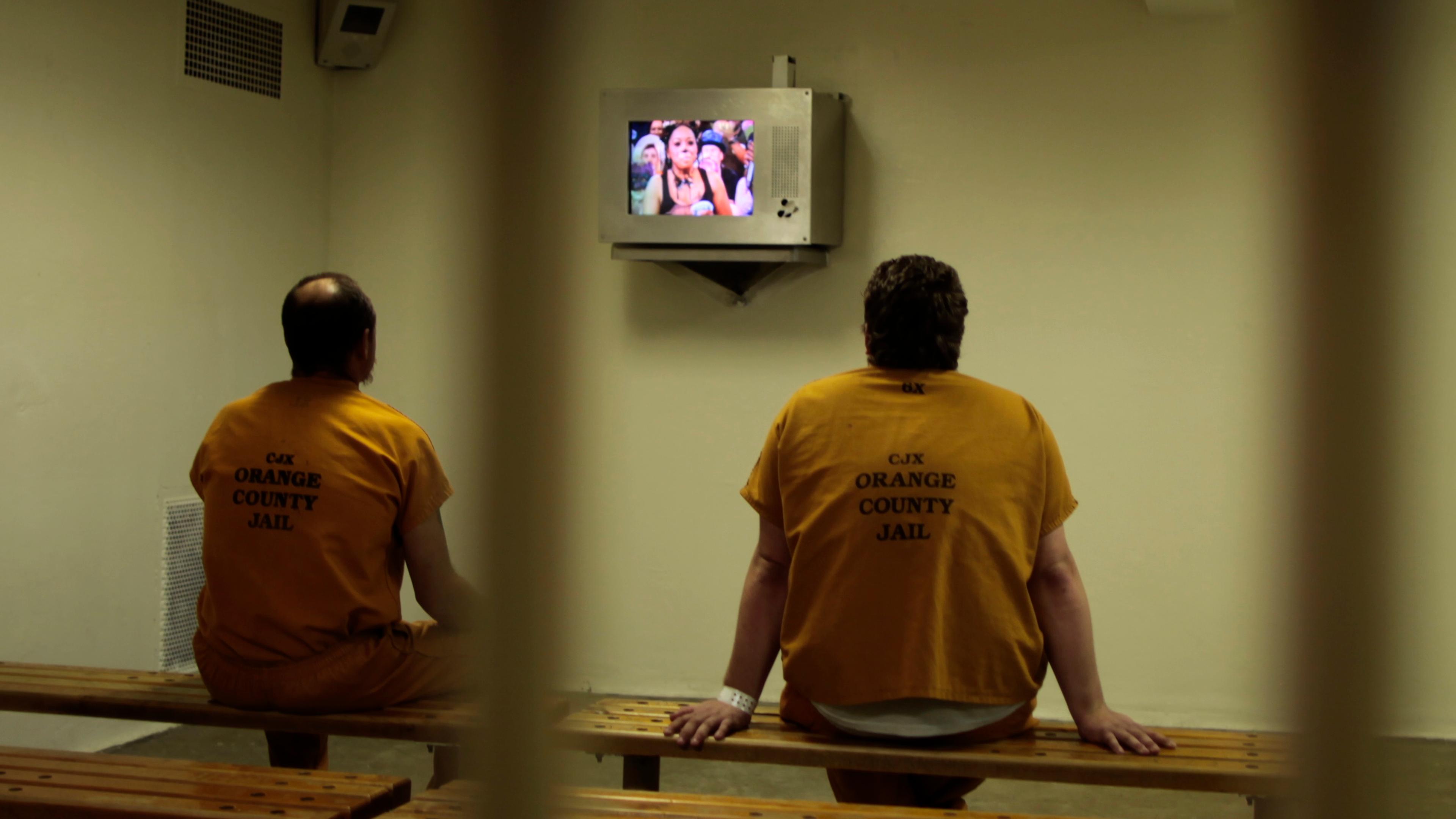 Photo of two inmates in orange uniforms sitting on benches watching a TV at Orange County Jail.