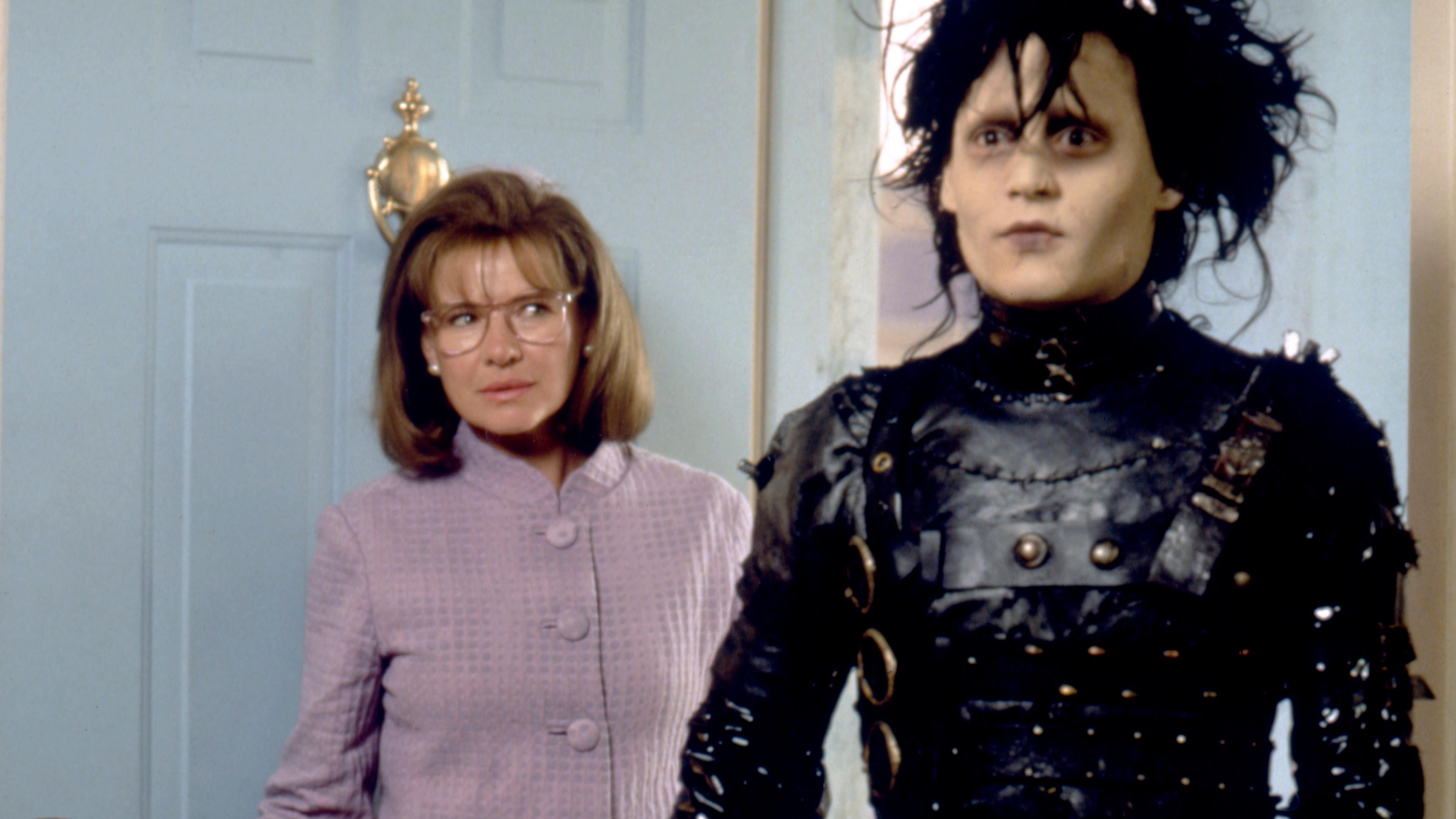 Scene from a film with a pale man in black leather and a woman in glasses standing by a blue door in a home setting.
