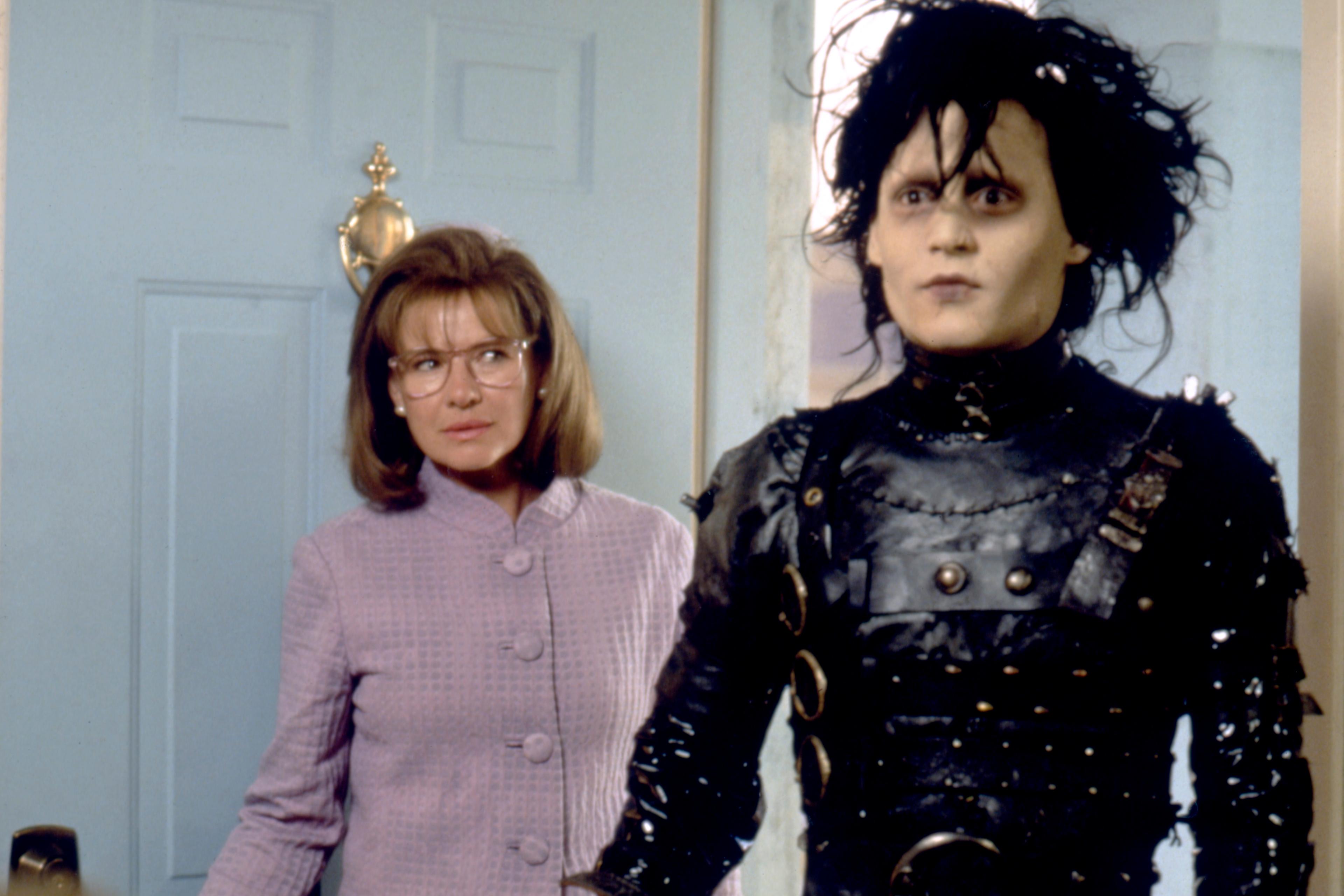 Scene from a film with a pale man in black leather and a woman in glasses standing by a blue door in a home setting.