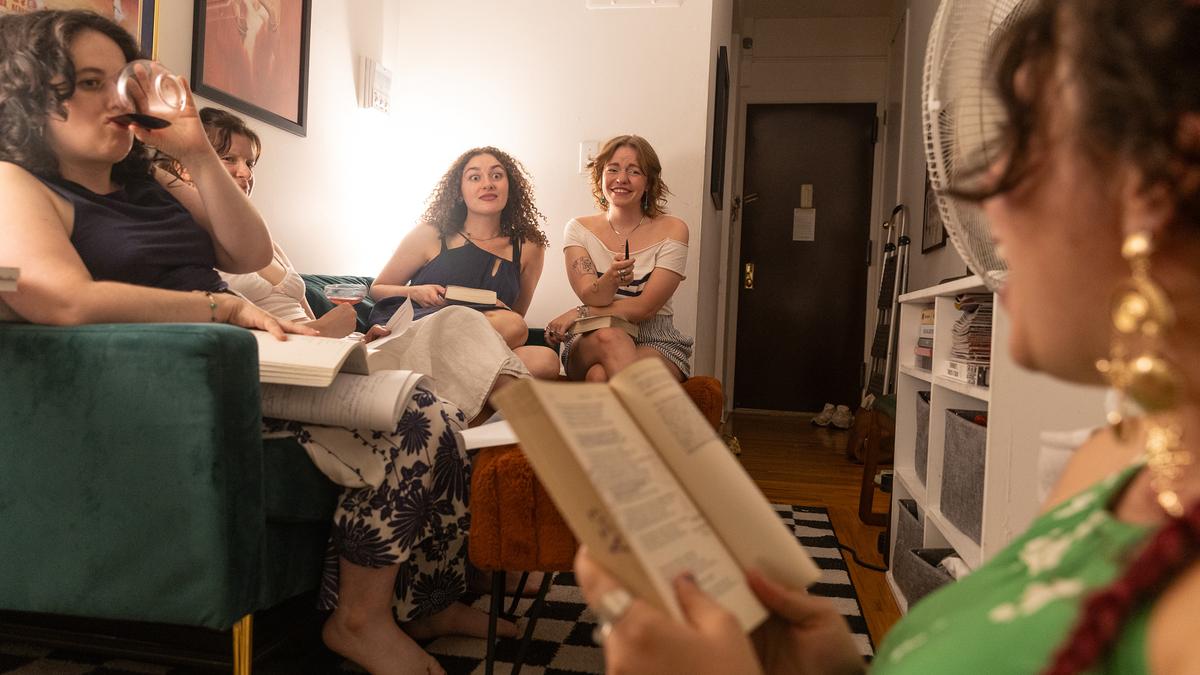 A book club meeting with five women, some reading, others chatting on a green sofa in a cosy living room.