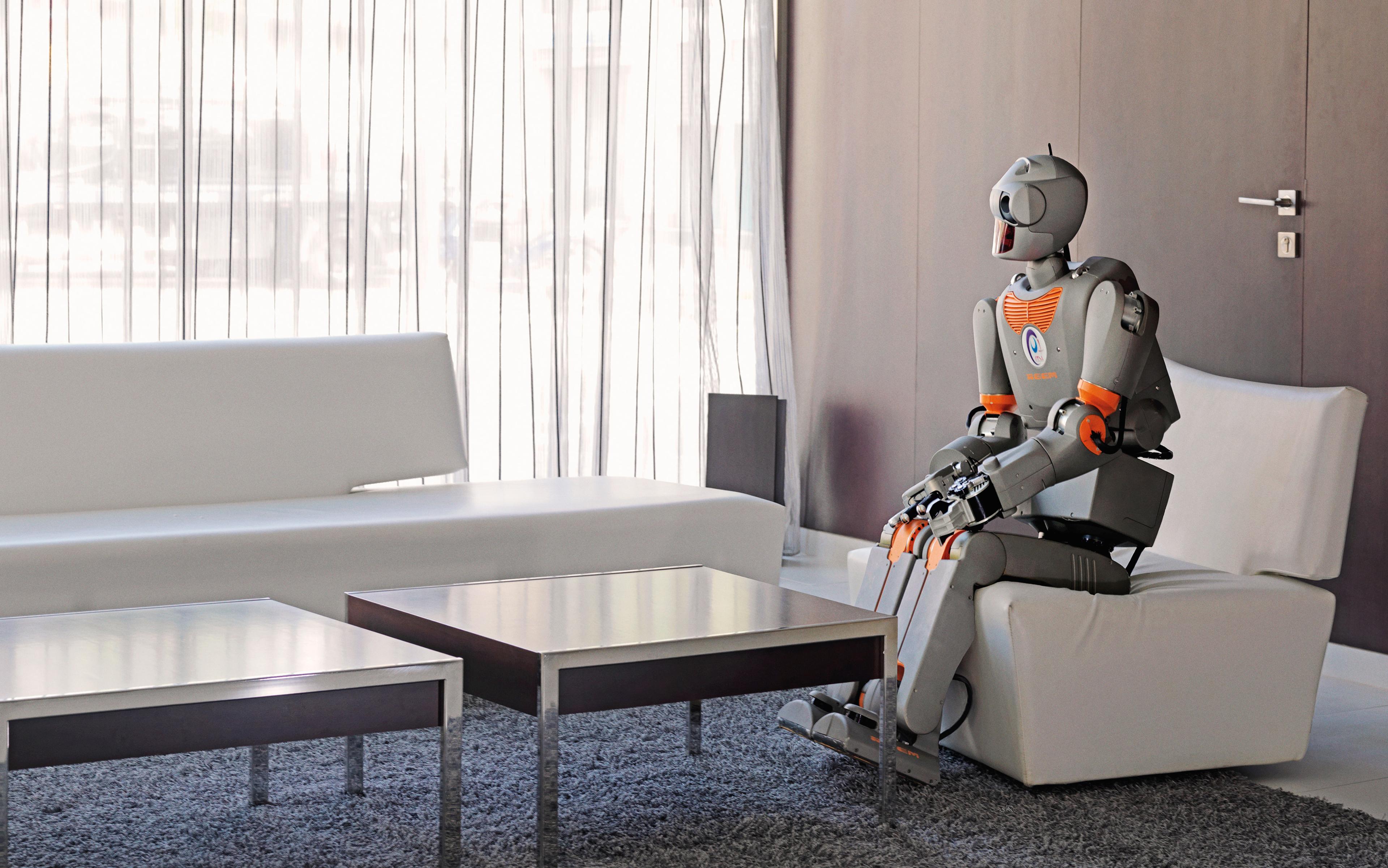 A robot sitting on a white chair in a modern living room with white furniture, a grey carpet and light curtains.