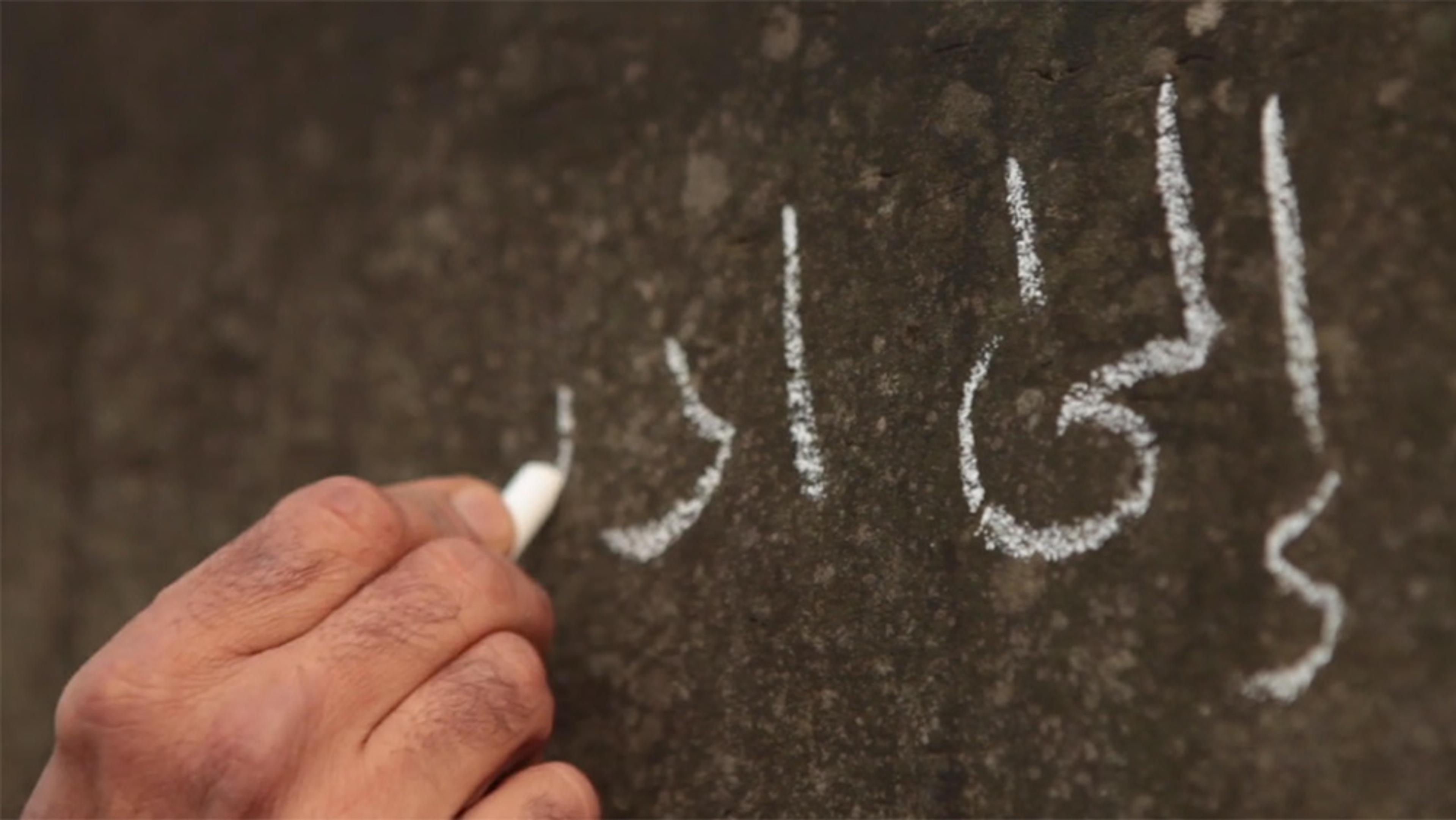 Close-up of a hand holding white chalk, writing Arabic script on a dark surface.