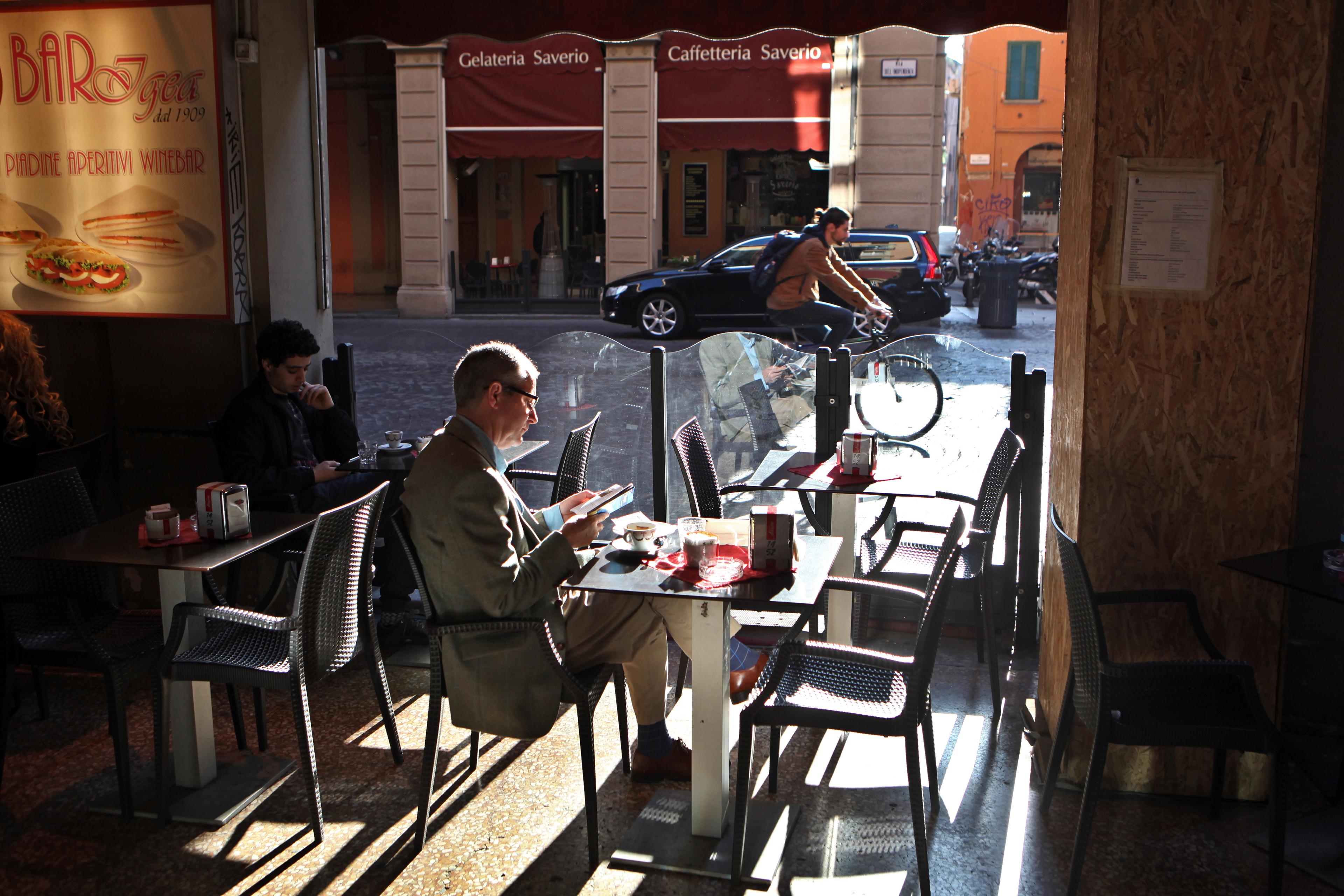Photo of a cafe interior with a man sitting alone reading, sunlight casting shadows others using phones, cyclist passing outside.