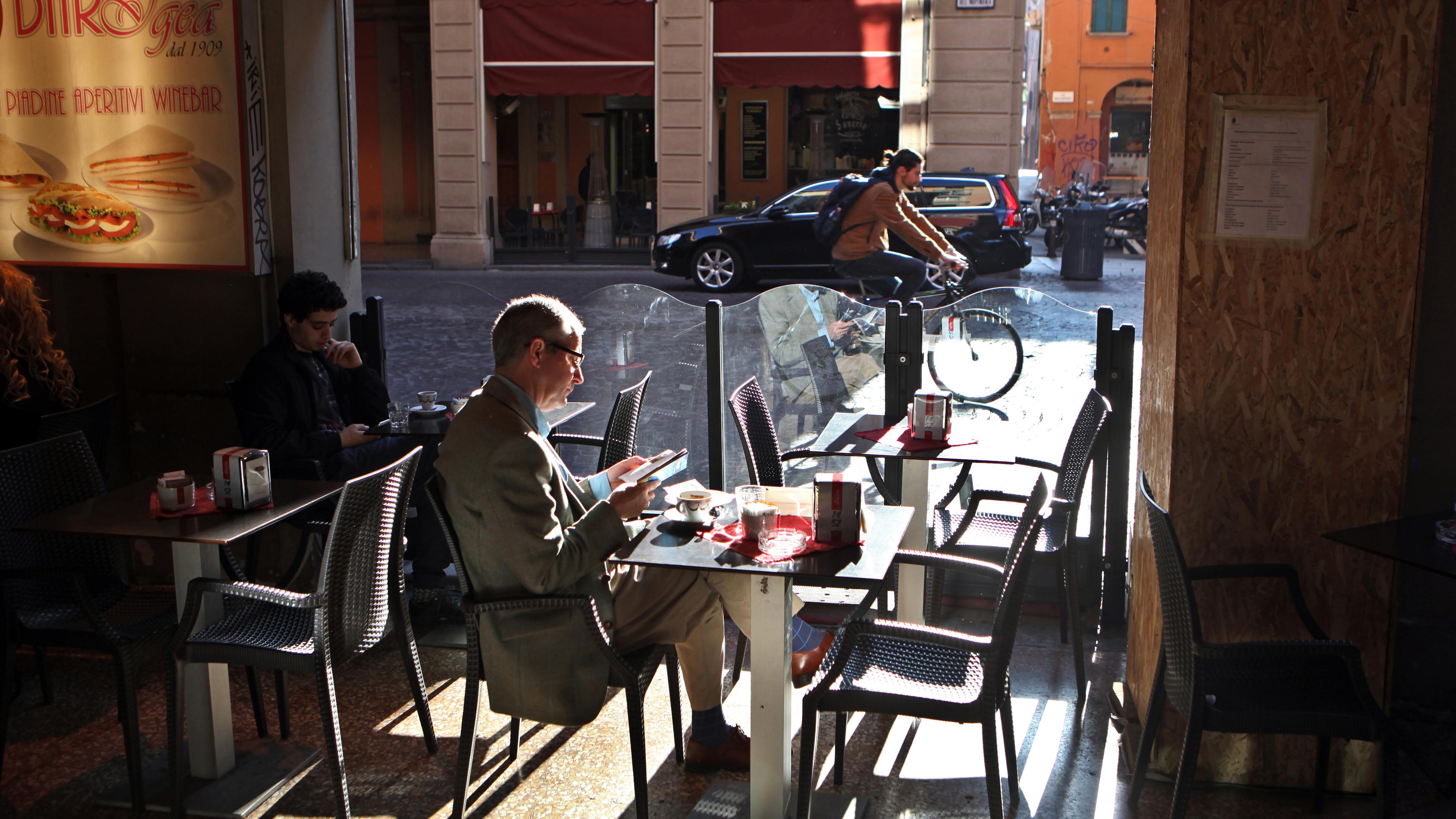 Photo of a cafe interior with a man sitting alone reading, sunlight casting shadows others using phones, cyclist passing outside.