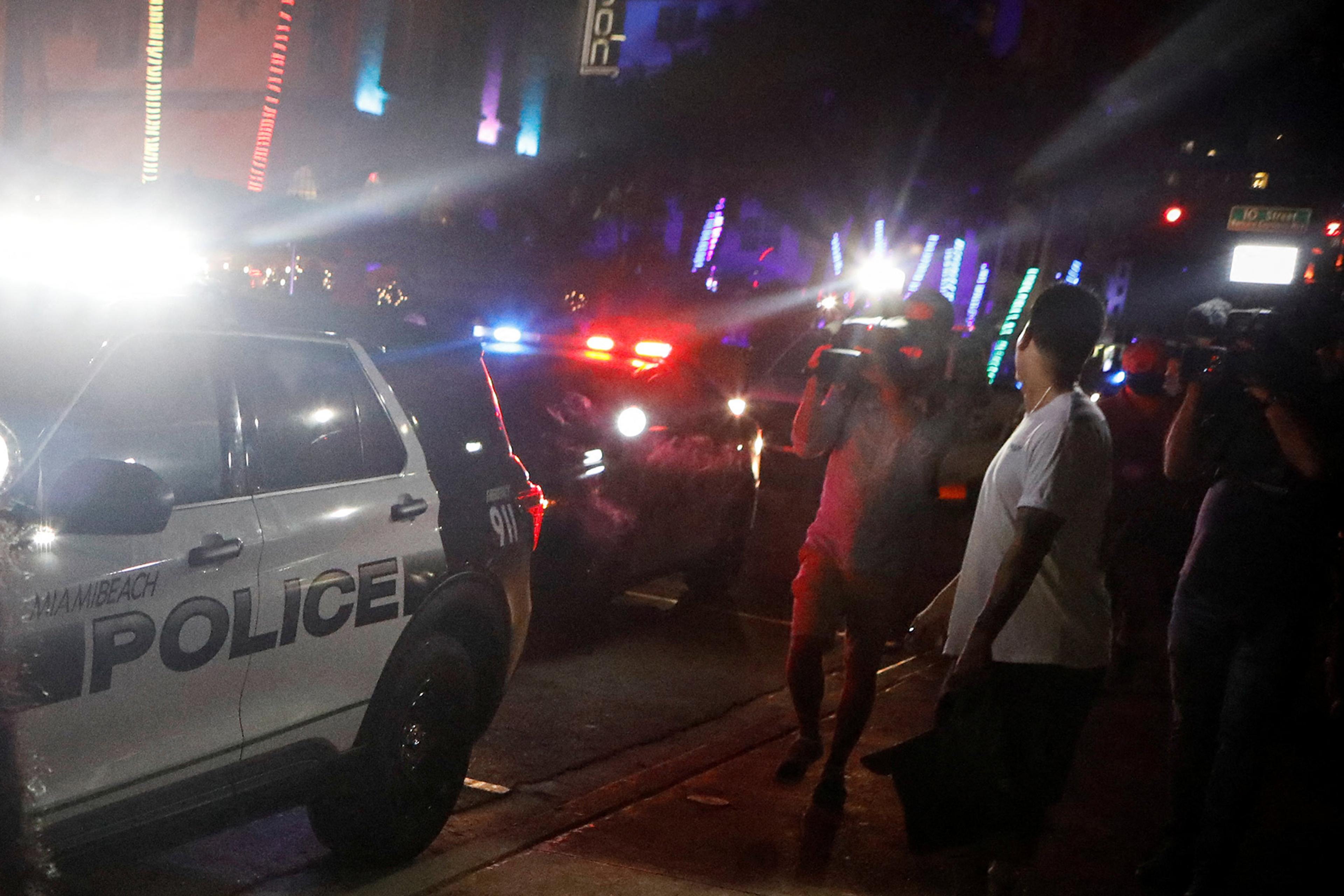 A Miami Beach police car with flashing lights surrounded by people and photographers at night.