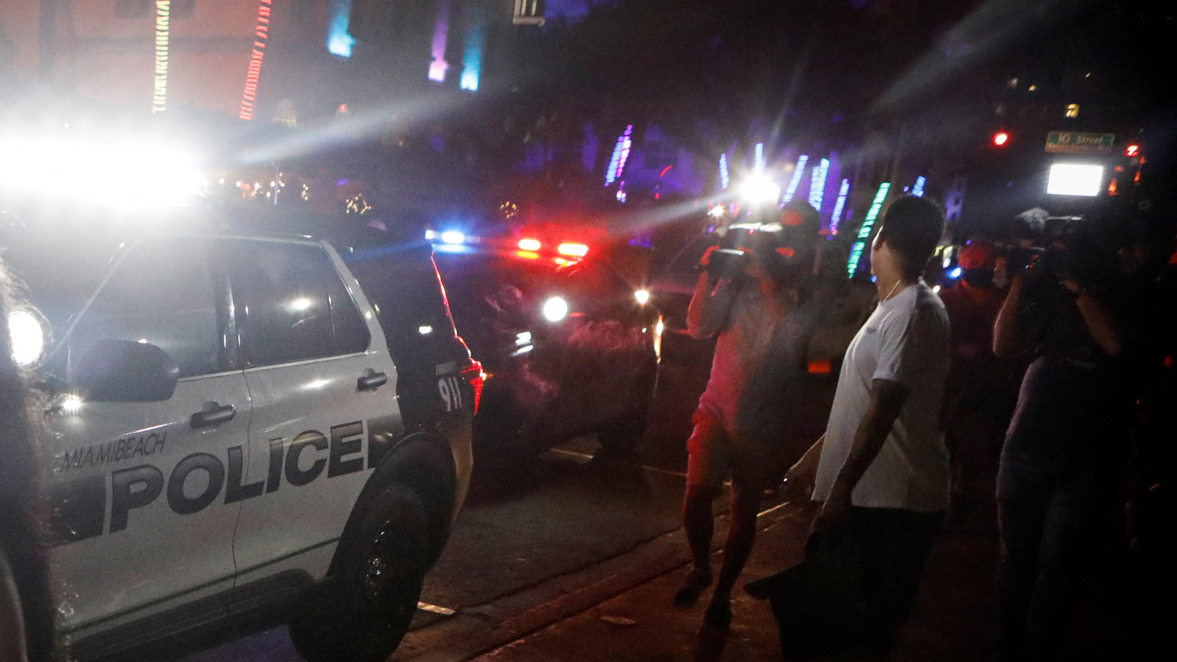 A Miami Beach police car with flashing lights surrounded by people and photographers at night.