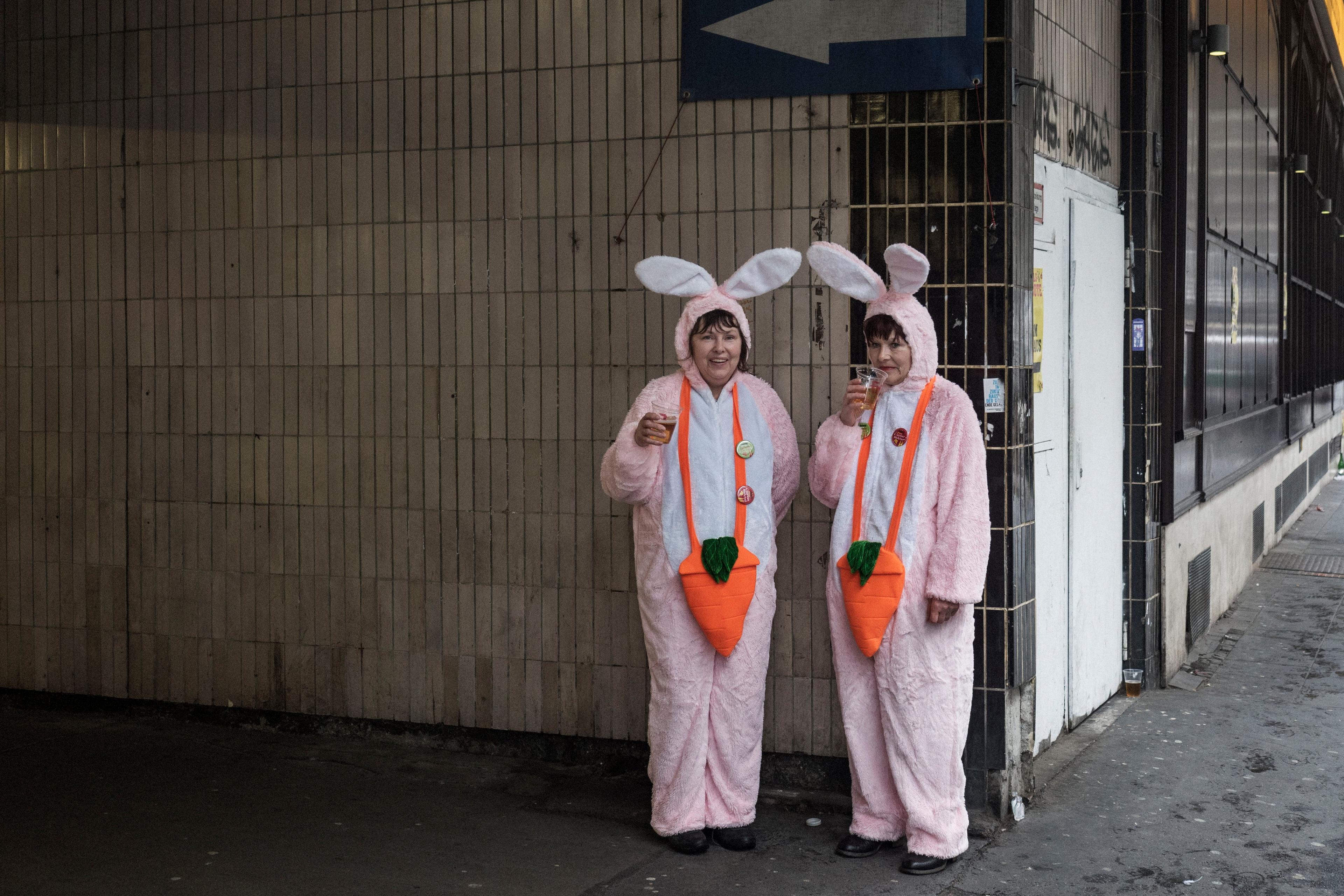 Photo of two people in pink bunny costumes with carrot designs, holding drinks and standing by a tiled wall under a sign.