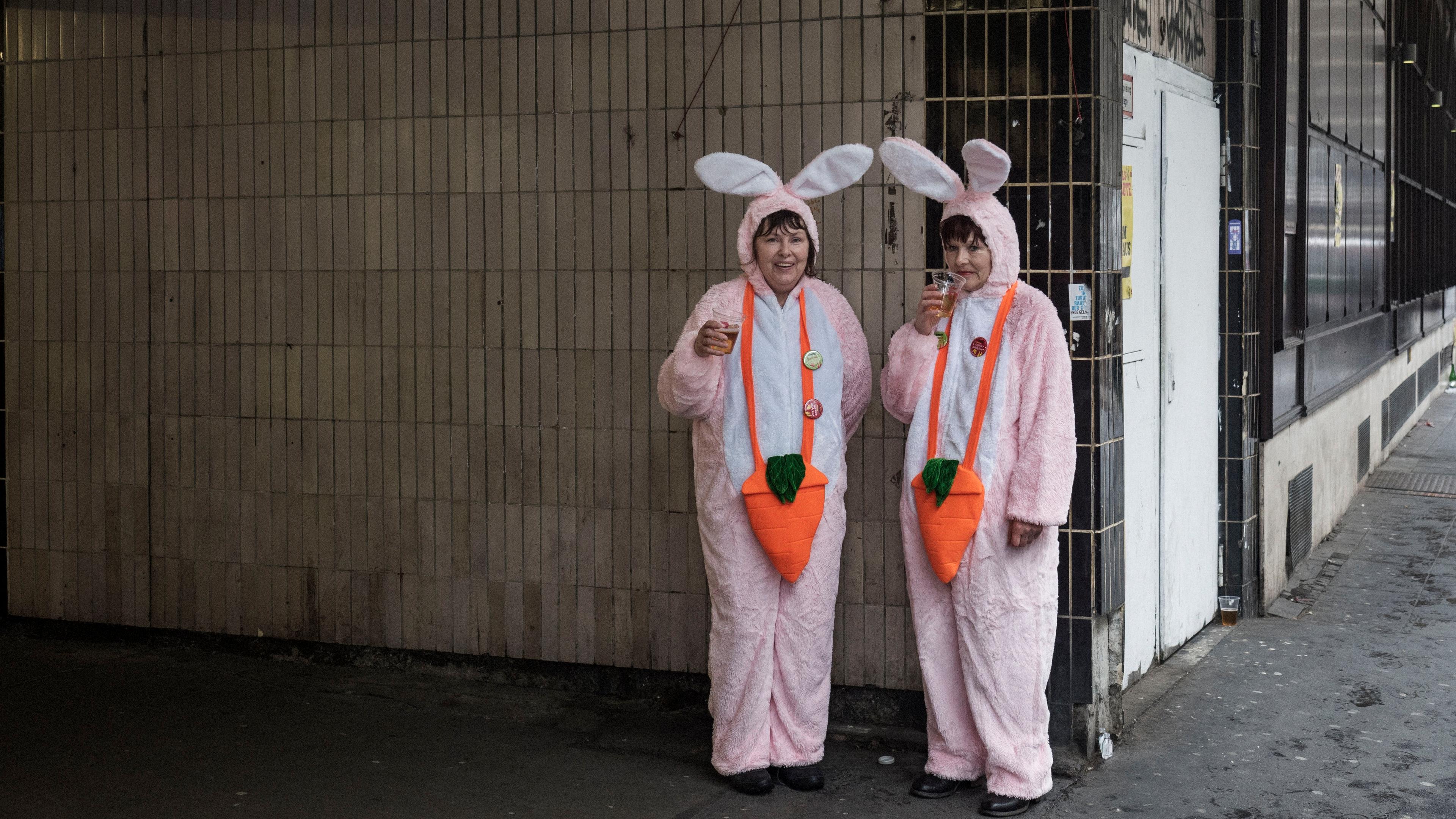 Photo of two people in pink bunny costumes with carrot designs, holding drinks and standing by a tiled wall under a sign.