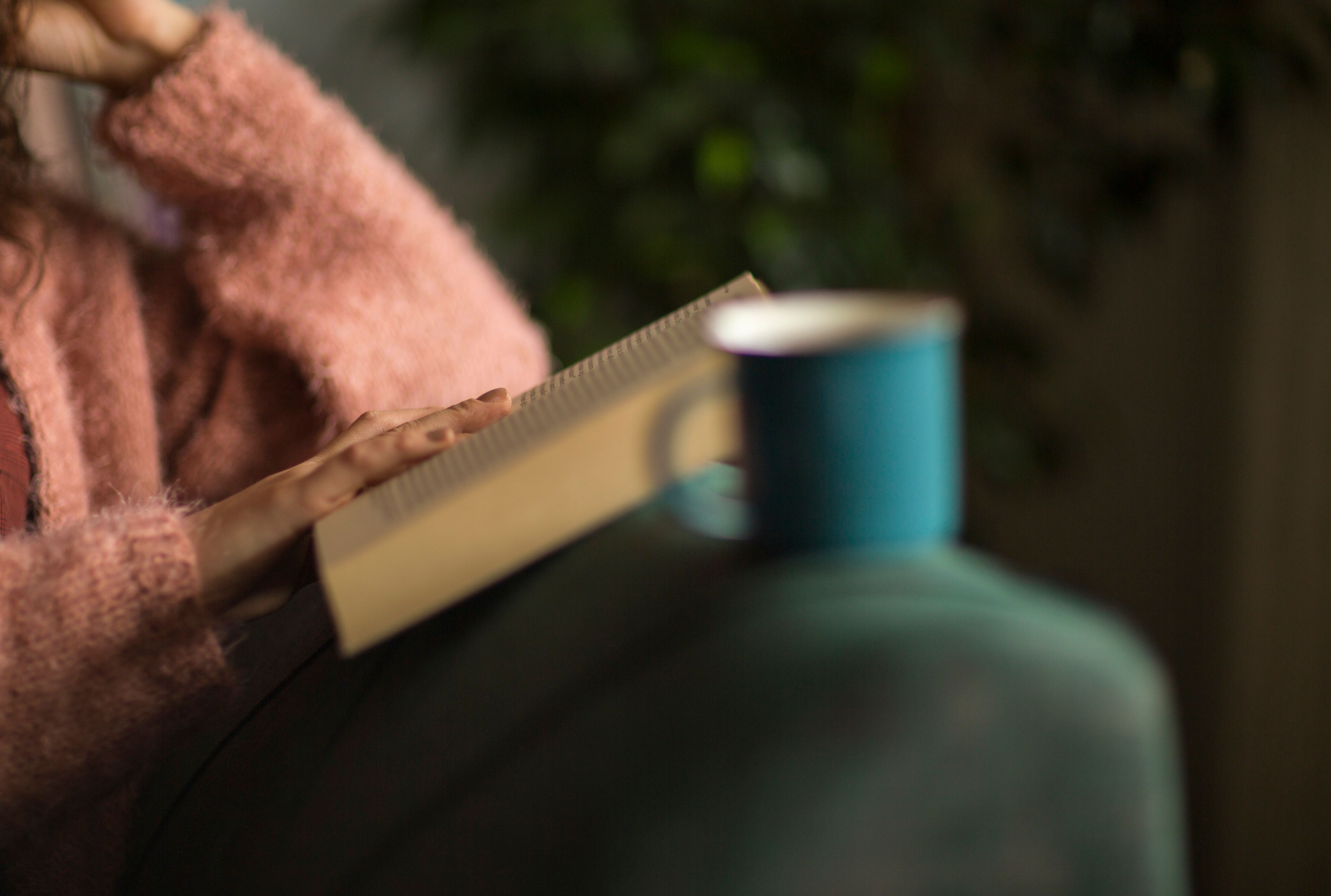 Photo of a person in a pink jumper reading a book on a sofa with a blue mug in the foreground.