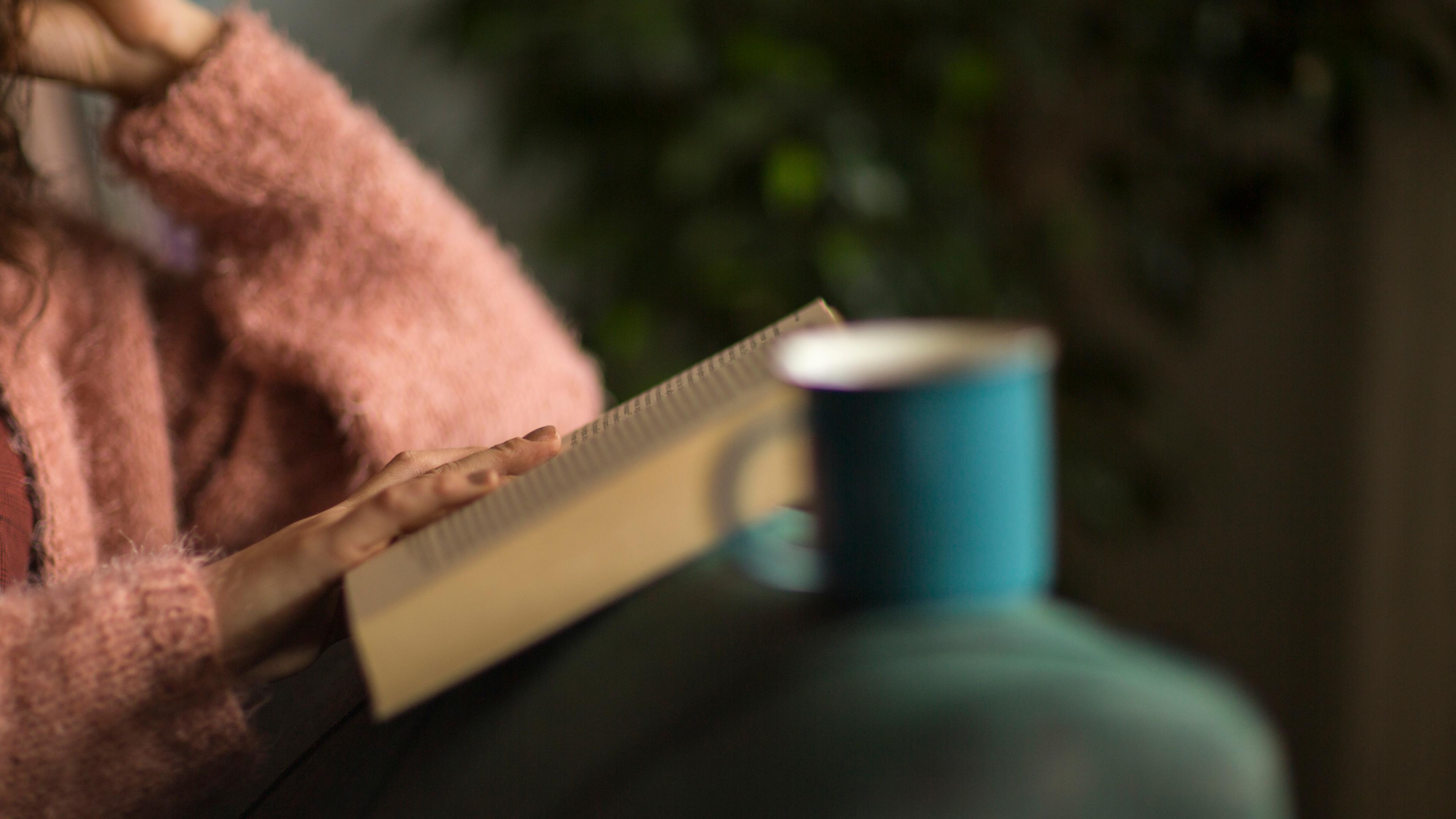 Photo of a person in a pink jumper reading a book on a sofa with a blue mug in the foreground.