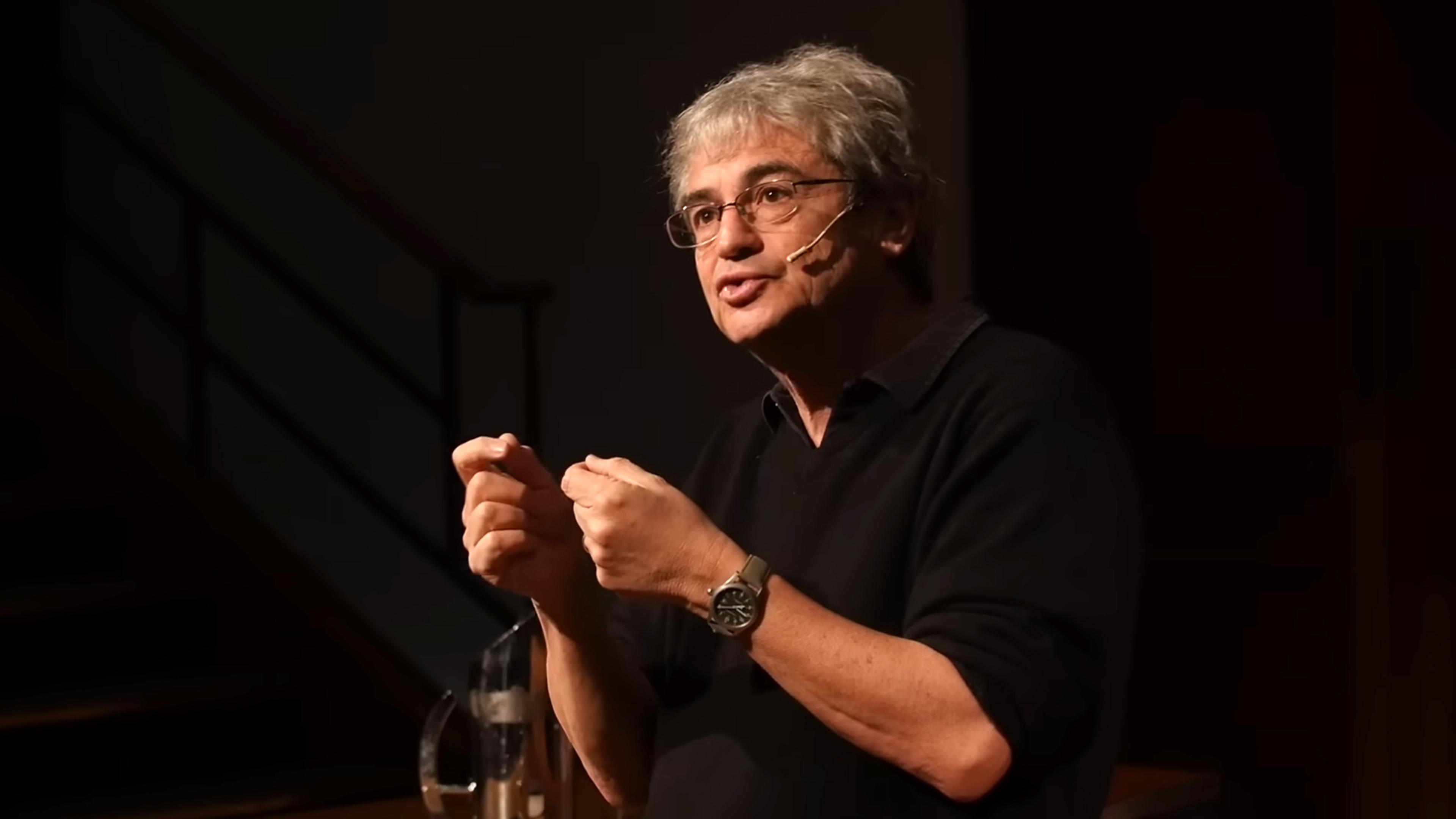 A man speaking on stage with a headset microphone wearing glasses and a black sweater gesturing expressively with his hands.