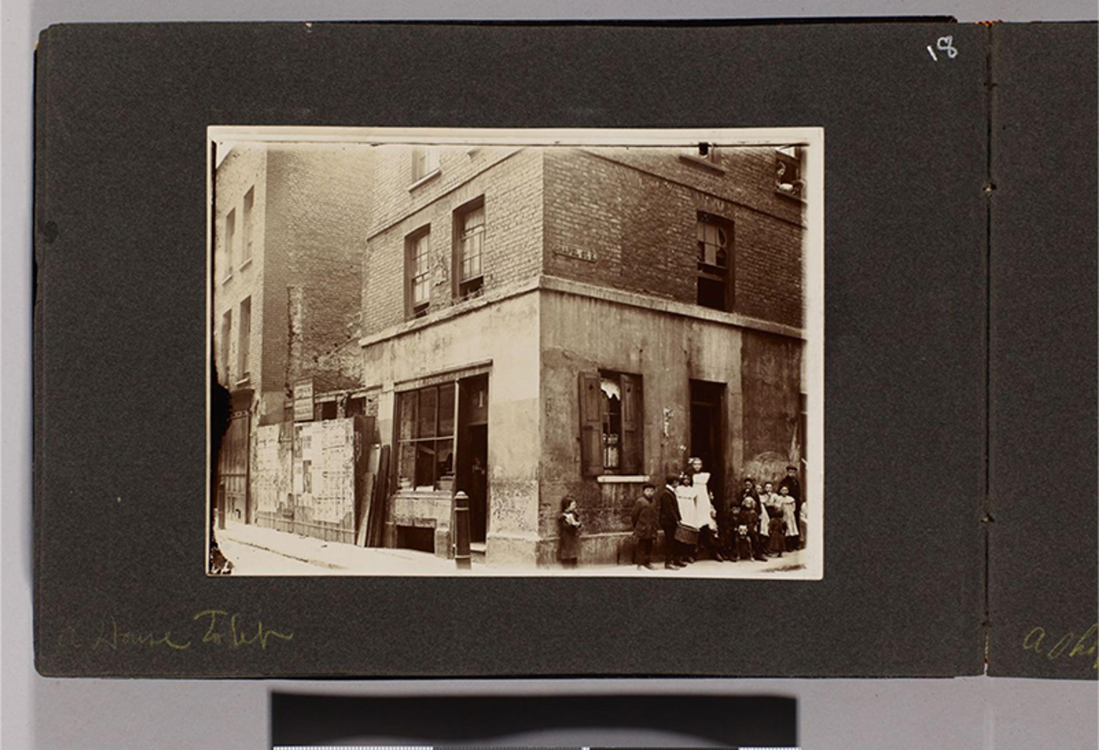 A sepia photo of a corner building with children and adults standing outside, the photo is mounted on a dark boarder.