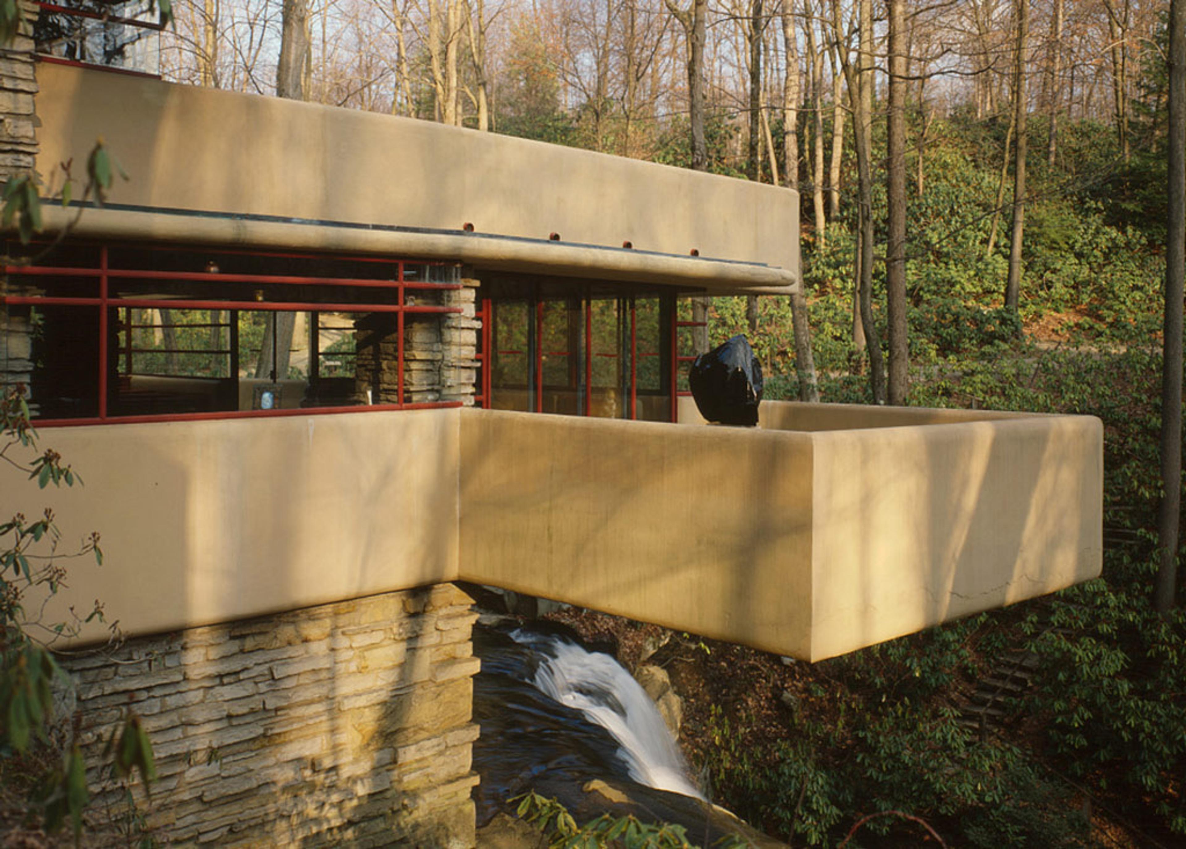 Photo of a modern house over a waterfall in a forest setting with trees and greenery surrounding the structure.