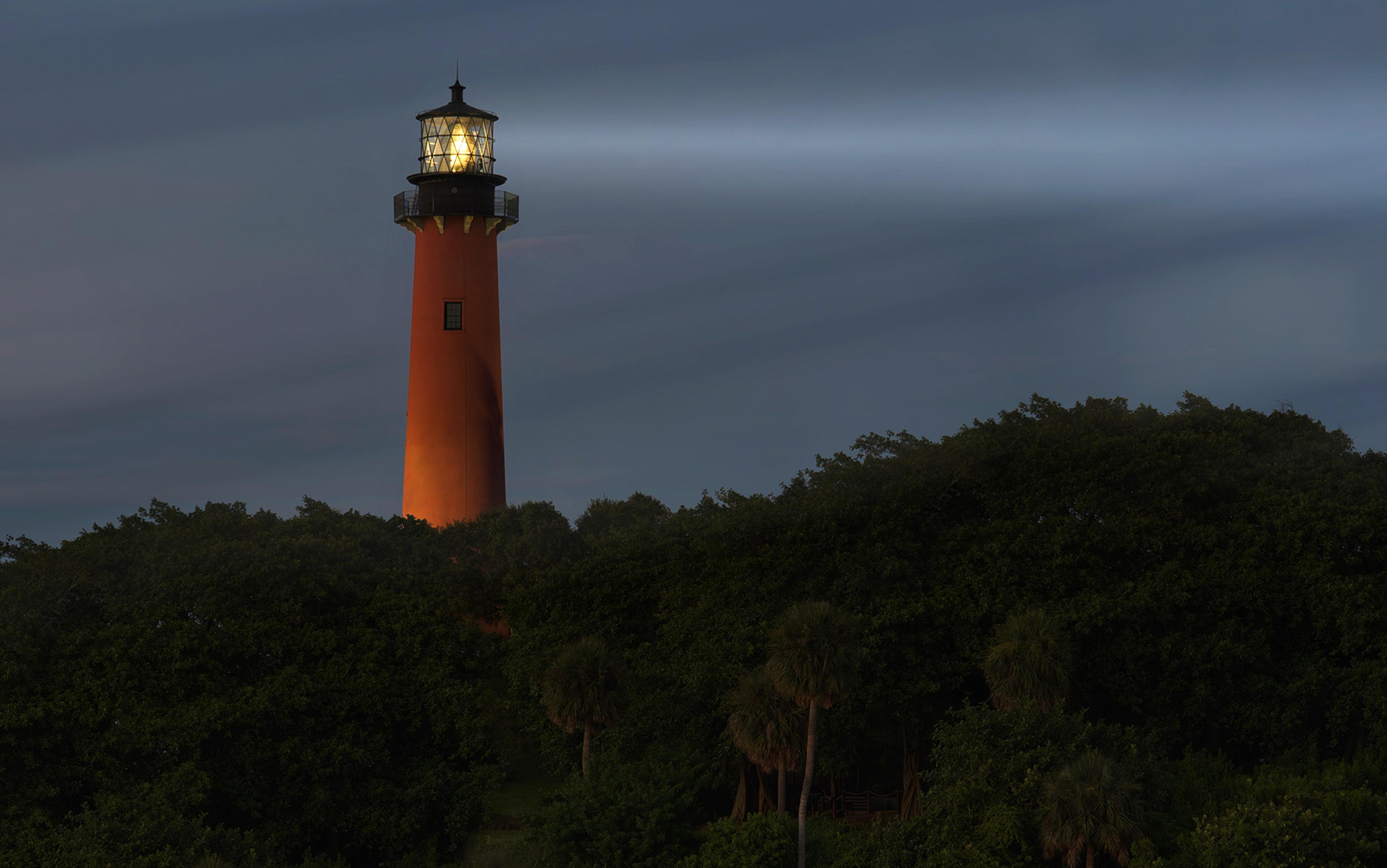 Illuminated lighthouse with beam of light, standing tall over dense green foliage during dusk.