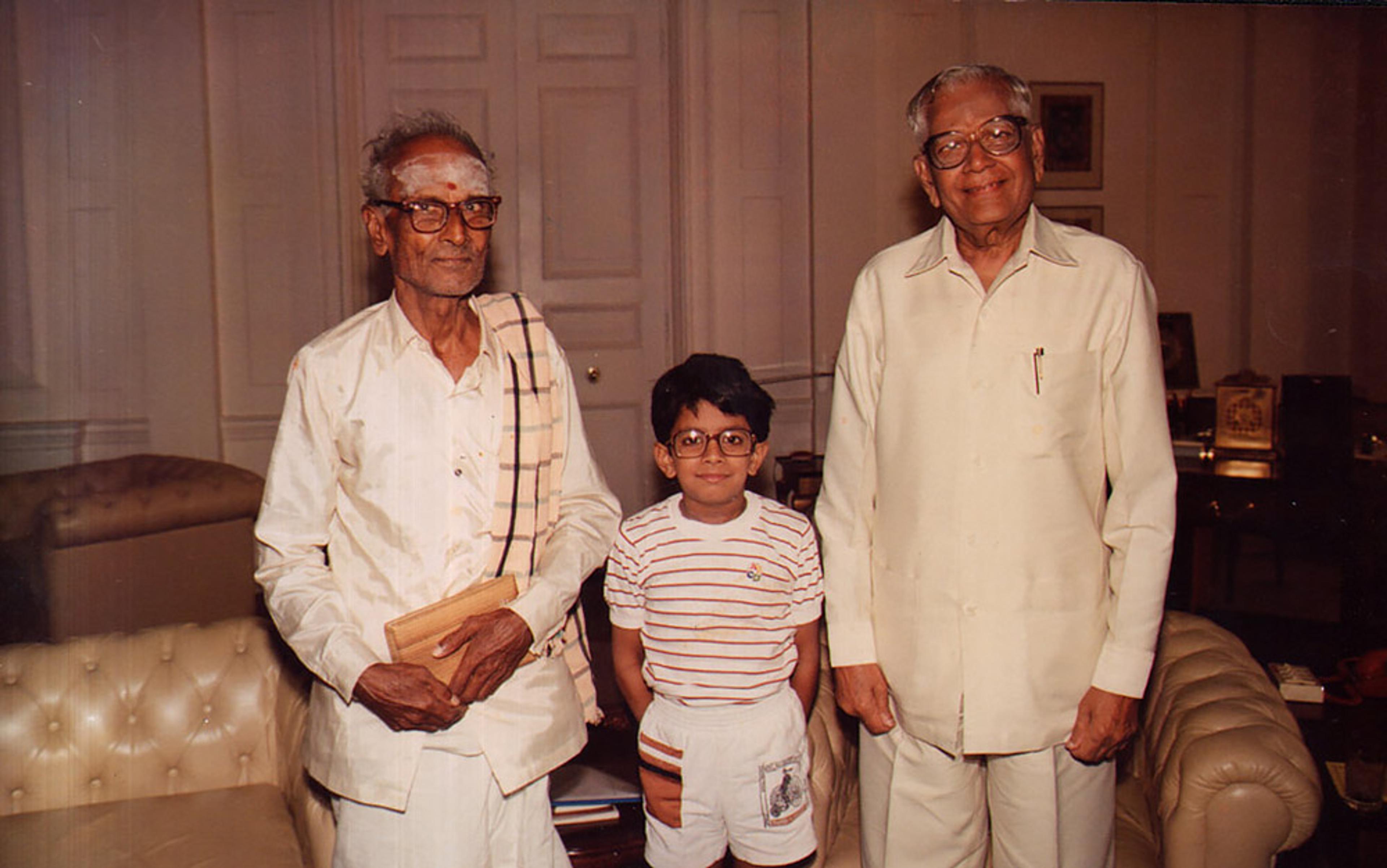 Two elderly men and a young boy standing indoors. They are smiling and dressed in casual clothing.