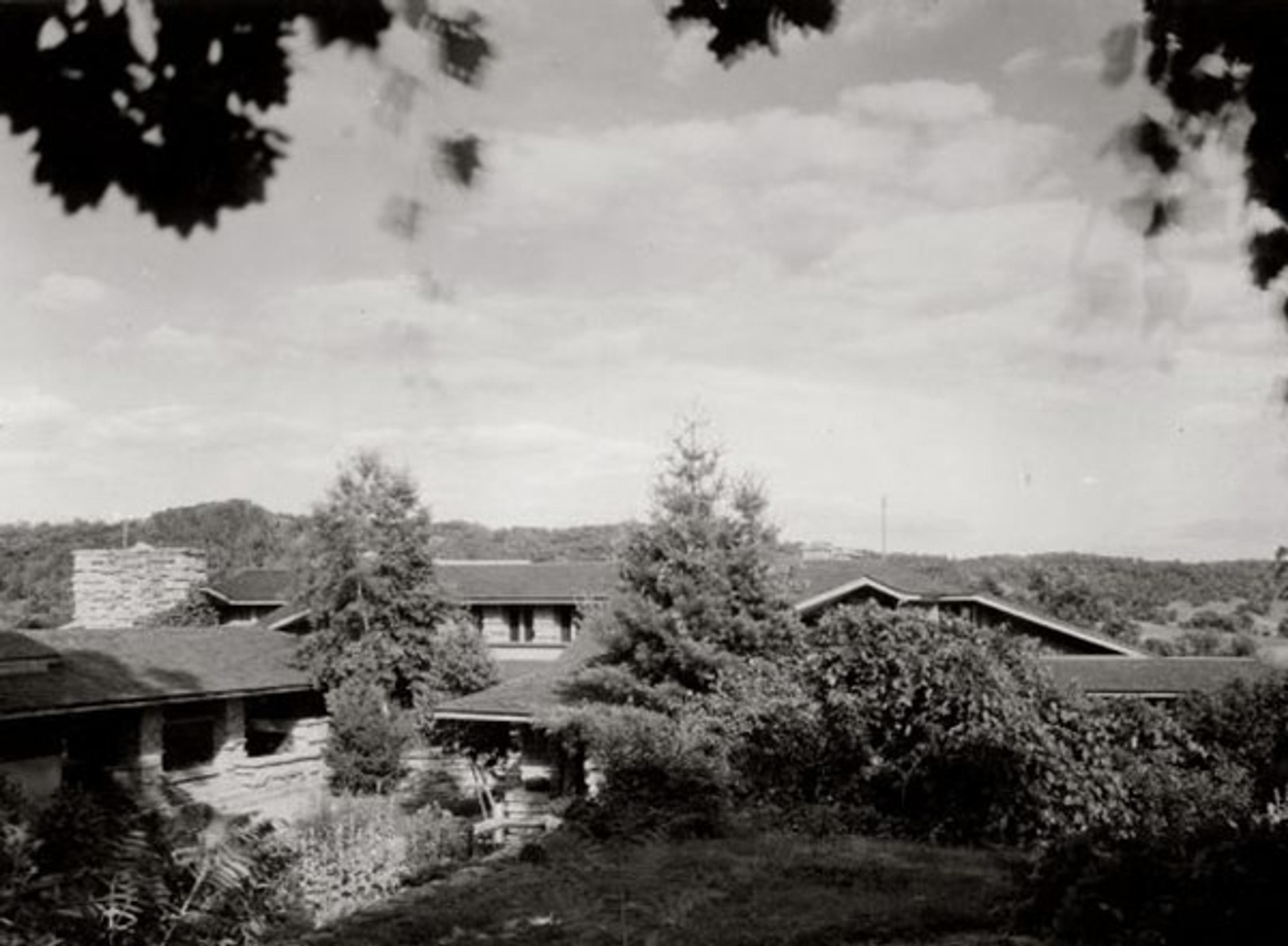 Black and white photo of a house surrounded by trees and shrubs with a cloudy sky in the background.