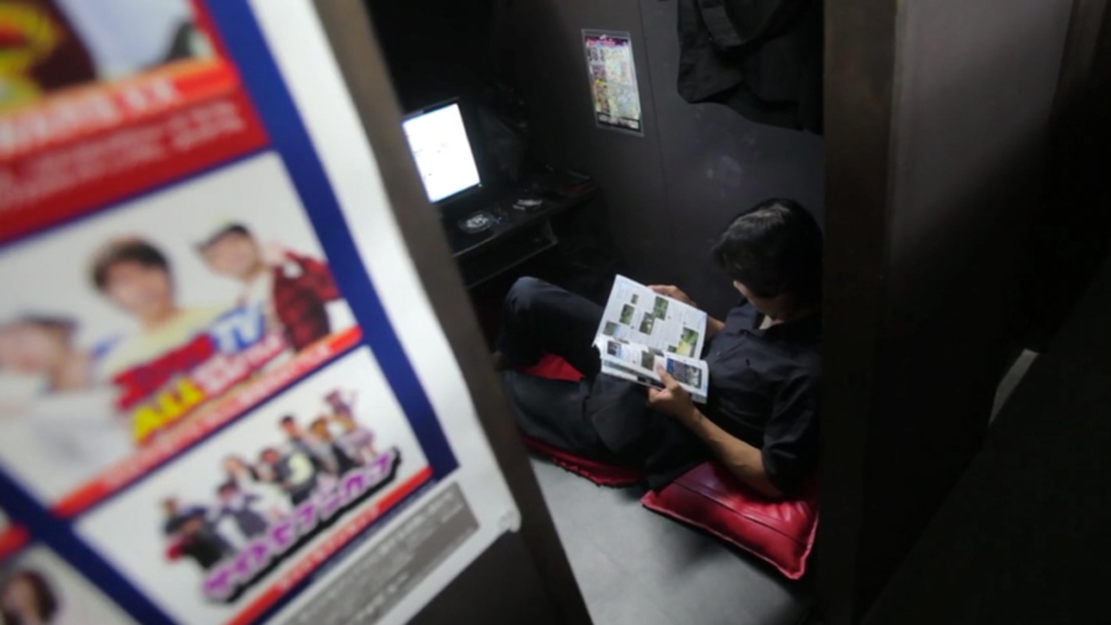 A man in a small booth reading a magazine with a computer screen in front of him and a poster on the door.