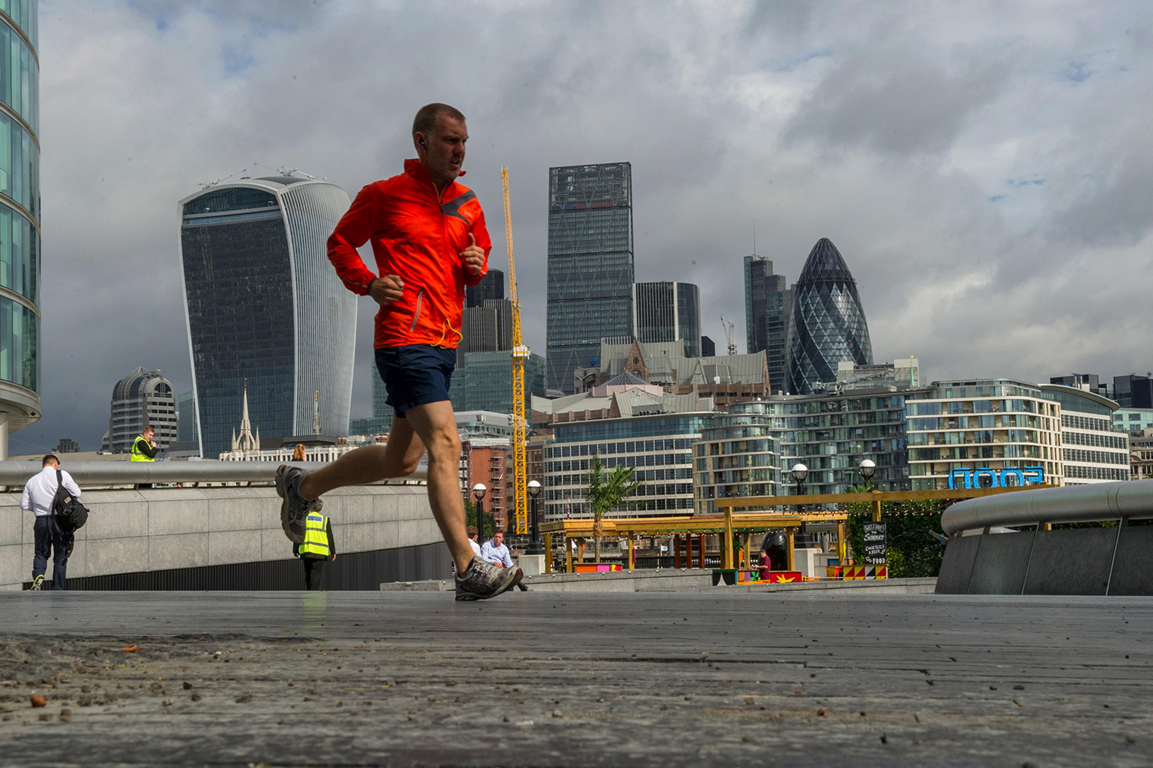A man in a red jacket jogs near iconic London skyscrapers, including the Gherkin and the Walkie Talkie, on a cloudy day. People walk in the background.