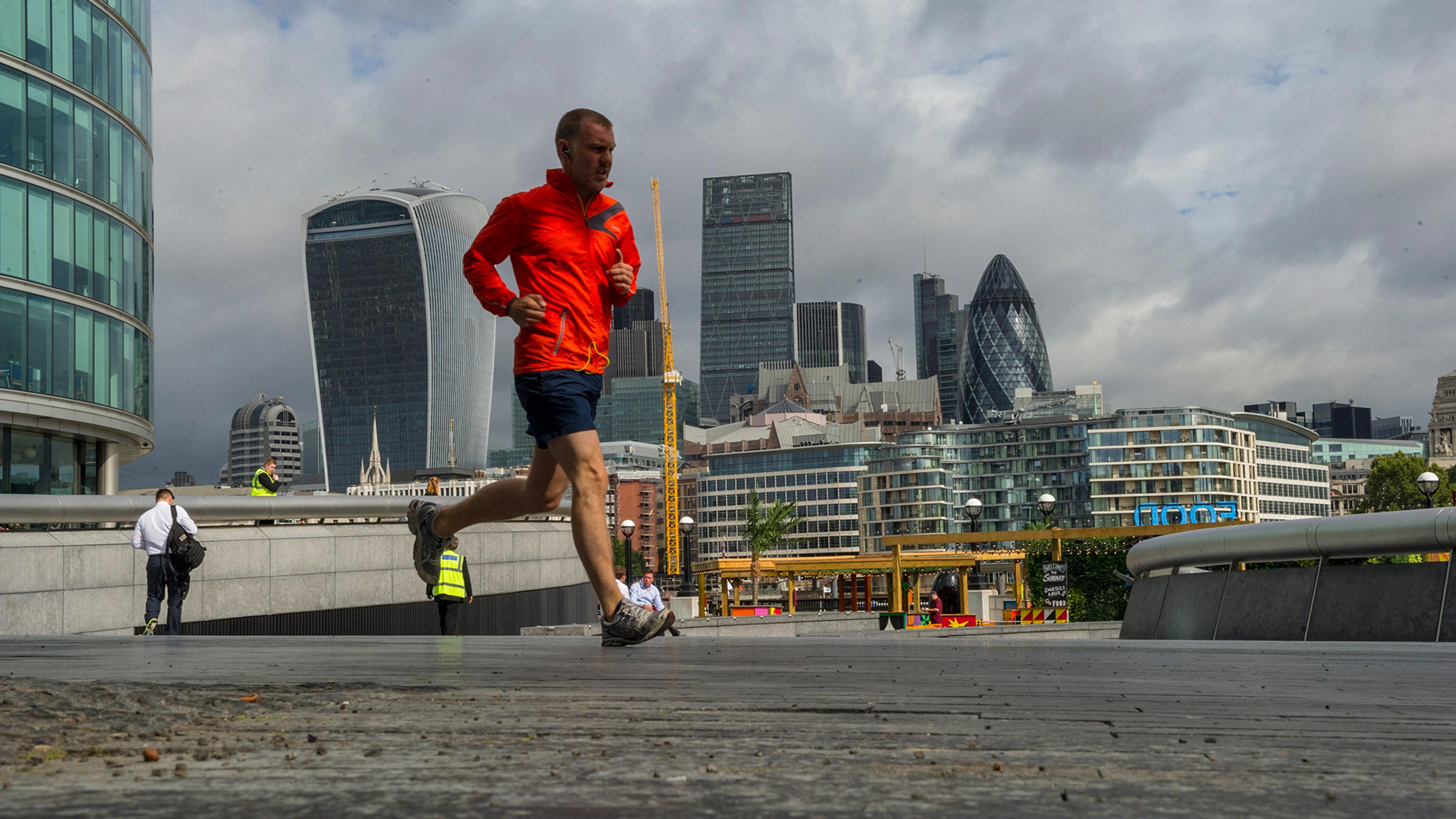 A man in a red jacket jogs near iconic London skyscrapers, including the Gherkin and the Walkie Talkie, on a cloudy day. People walk in the background.