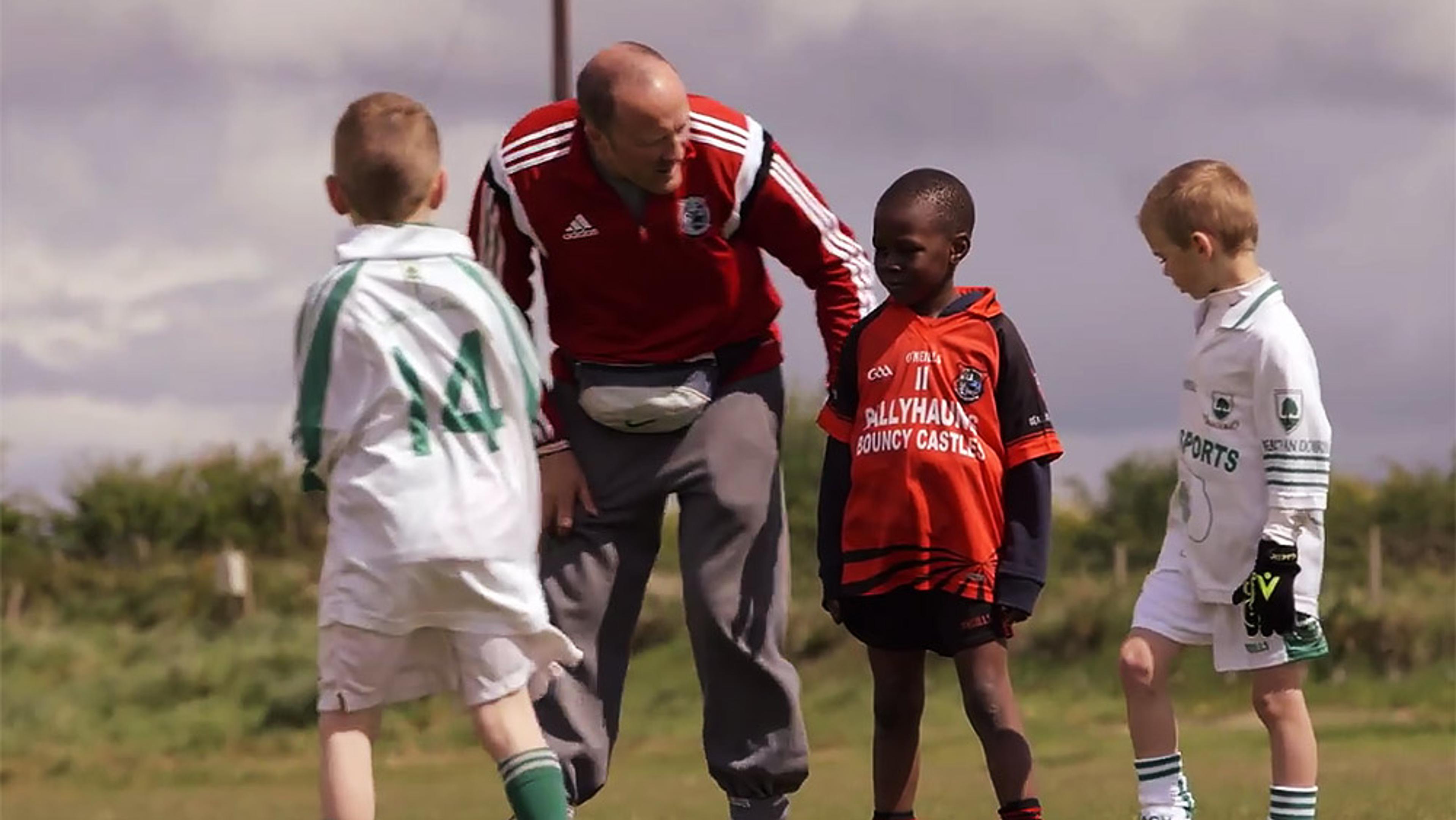 Photo of a coach in red speaking to three young boys in football kits on a grass field under a cloudy sky.
