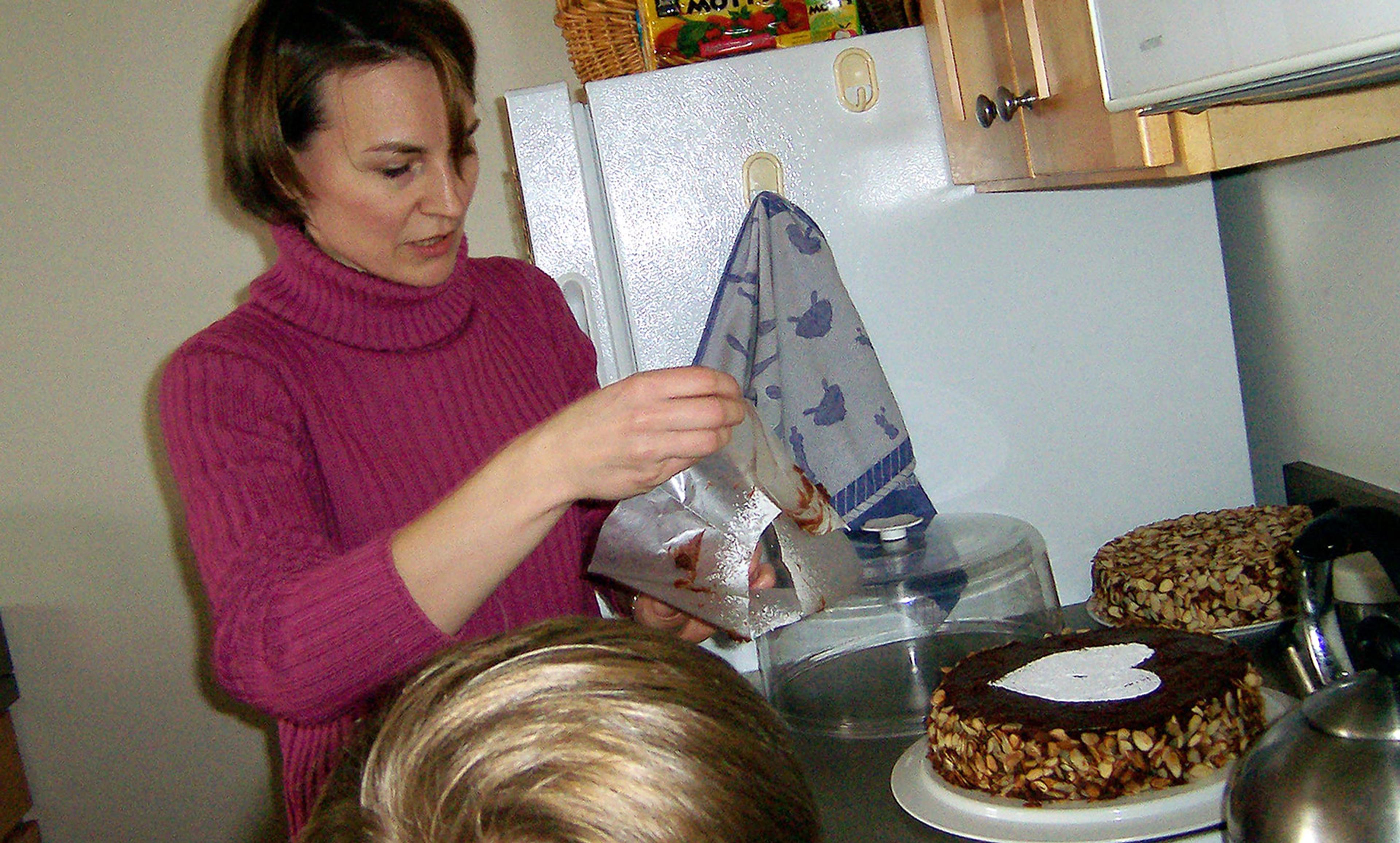A woman in a pink jumper decorating a cake in a kitchen with a fridge, two cakes, and a child’s head in the foreground.
