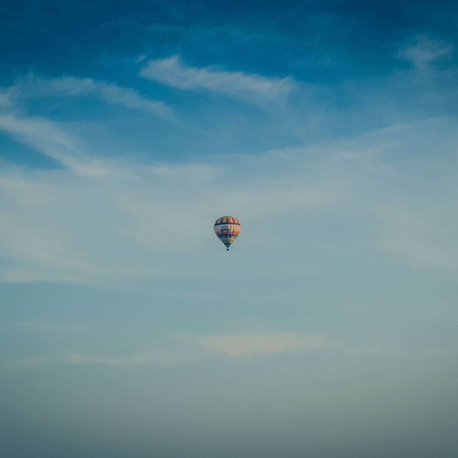 Photo of a single hot air balloon in a clear blue sky with light clouds.