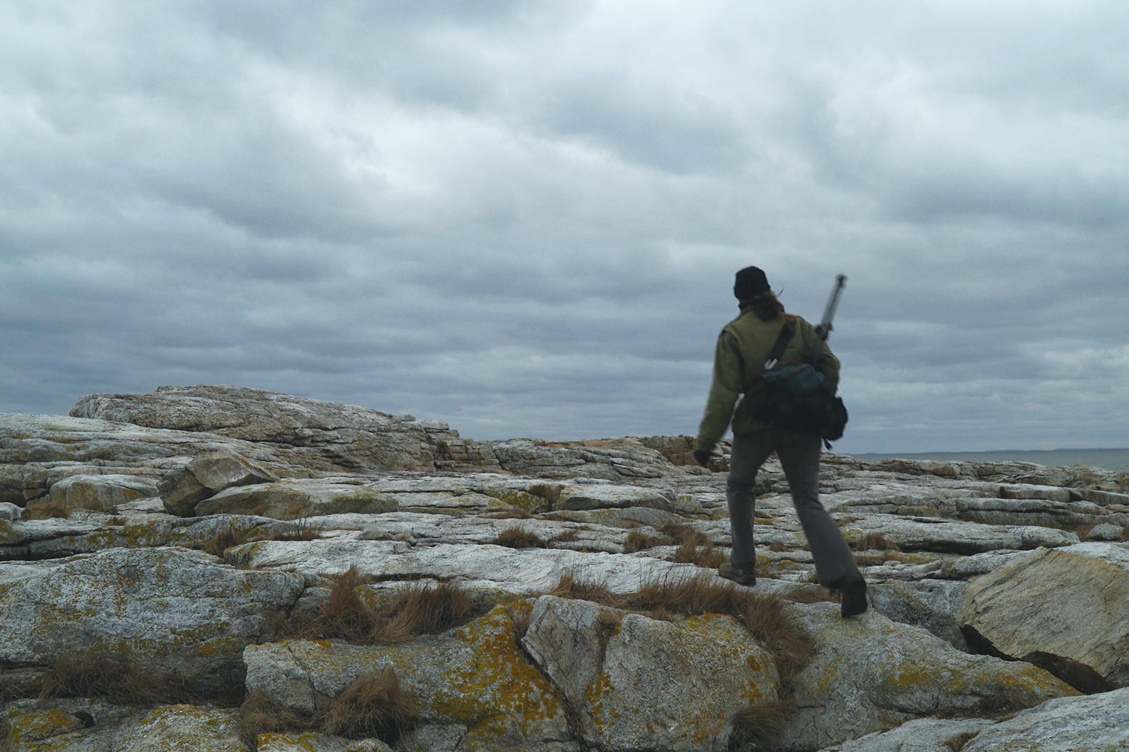 Person walking on a rocky landscape under a cloudy sky, carrying a backpack and a tool or instrument over their shoulder.