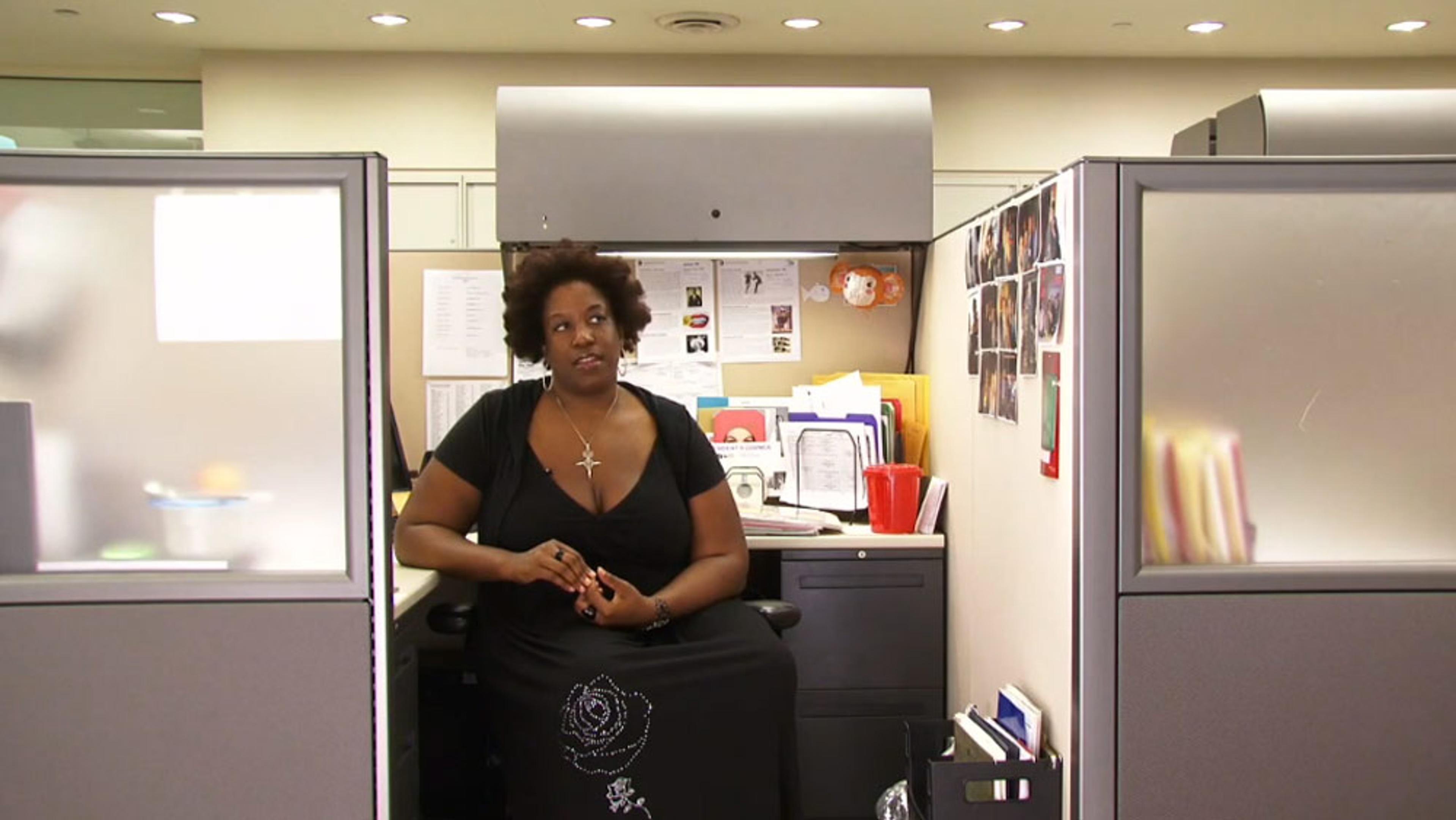 An office cubicle with a woman sitting on a chair, surrounded by papers, photos, and office supplies on her desk.