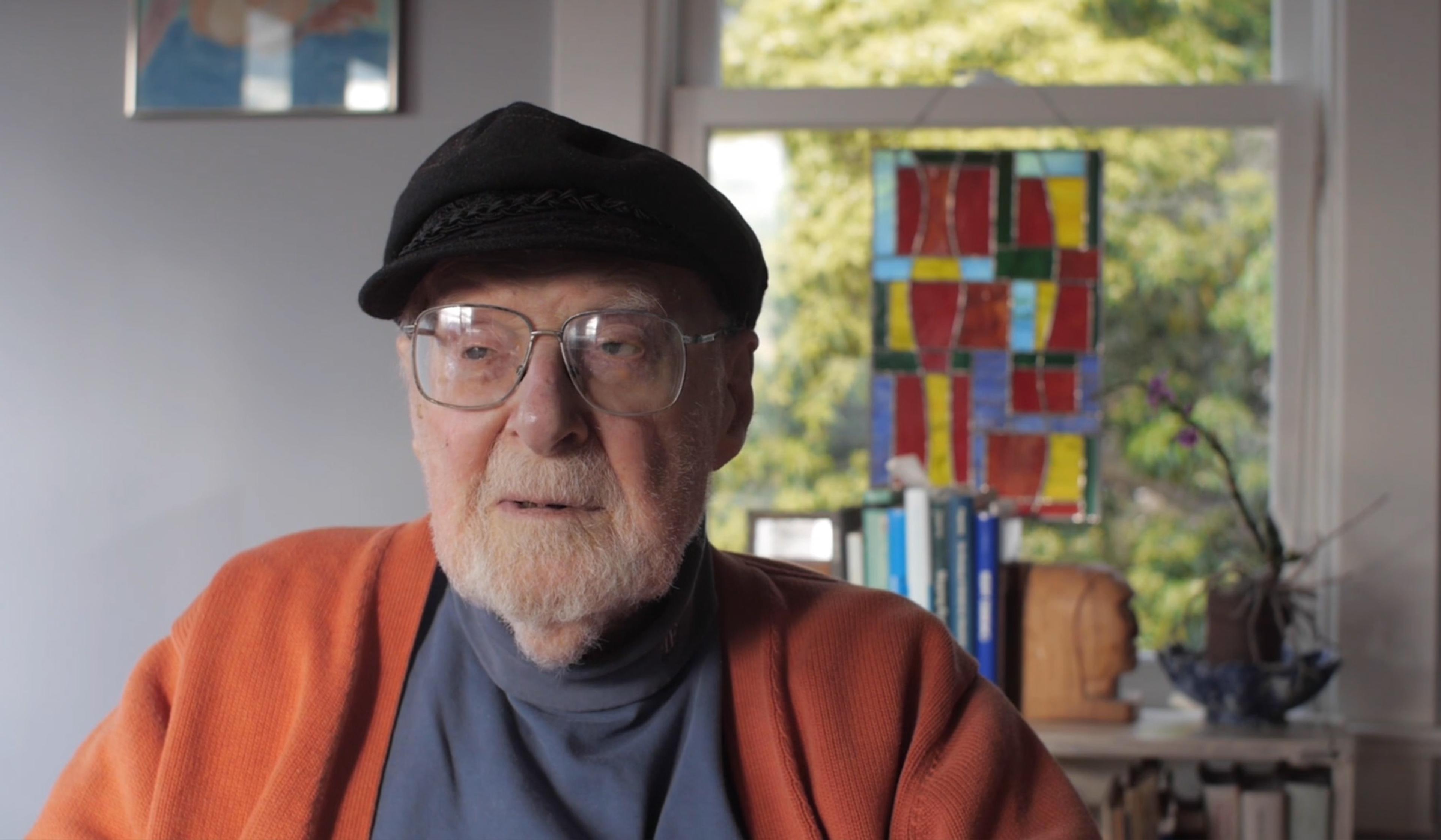Elderly man wearing glasses, a black hat and an orange cardigan, sitting indoors with a colourful stained glass window behind him.