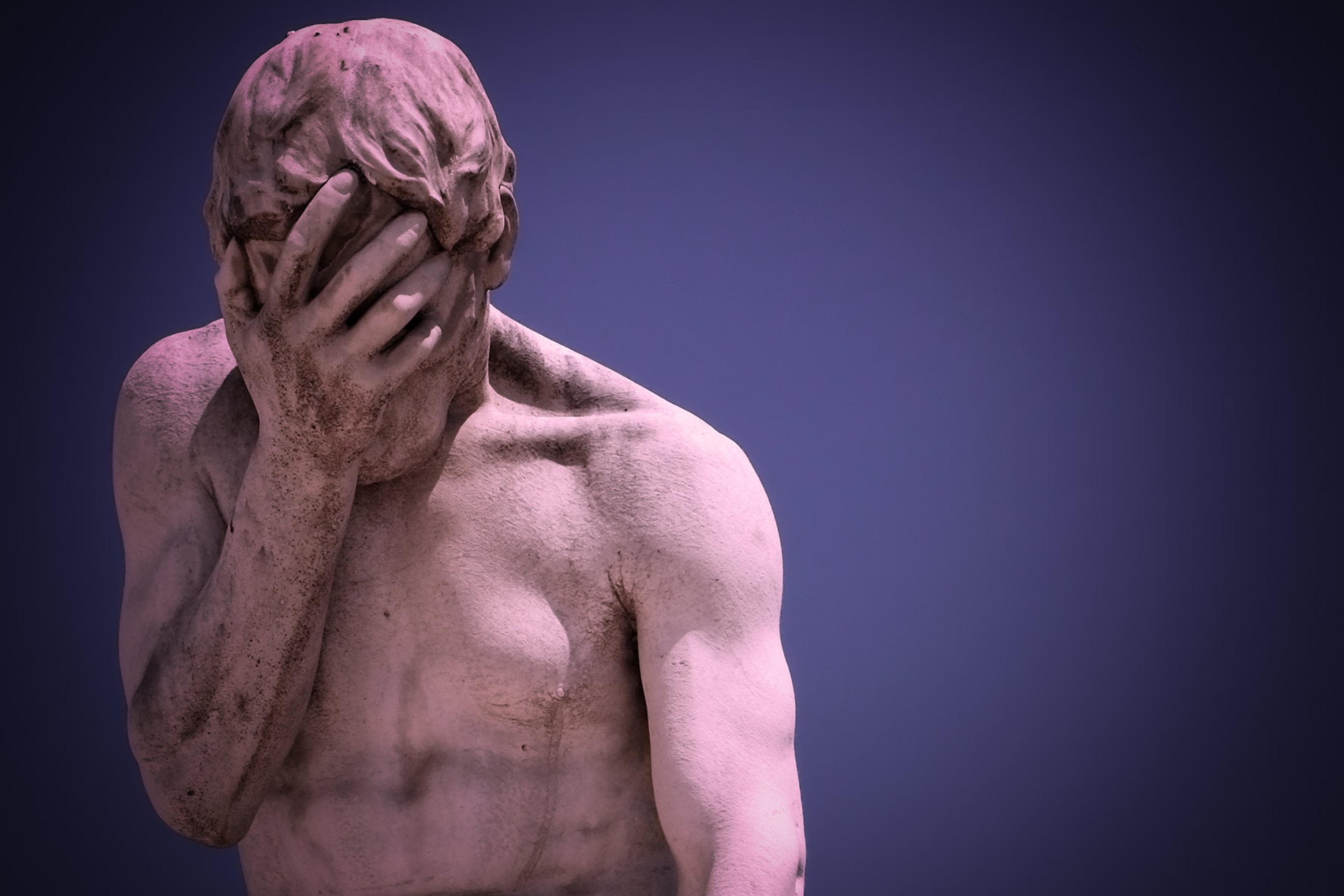 A marble statue of a man covering his face with his hand set against a dark blue background.