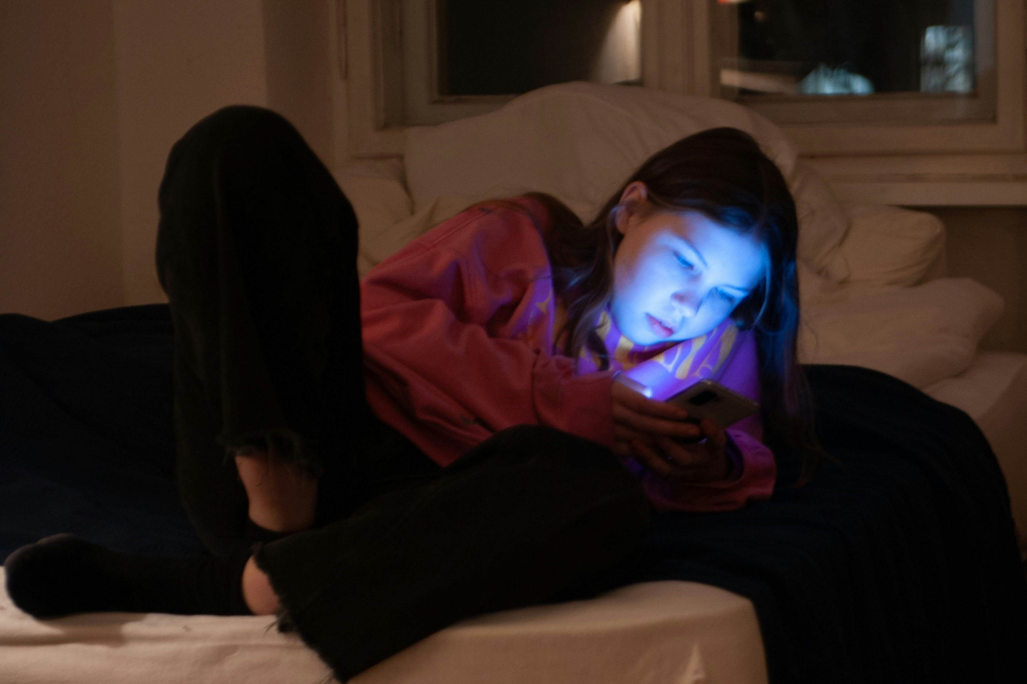 A young girl in a pink hoodie lit by a phone screen, lying on a bed in a dimly lit room at night.