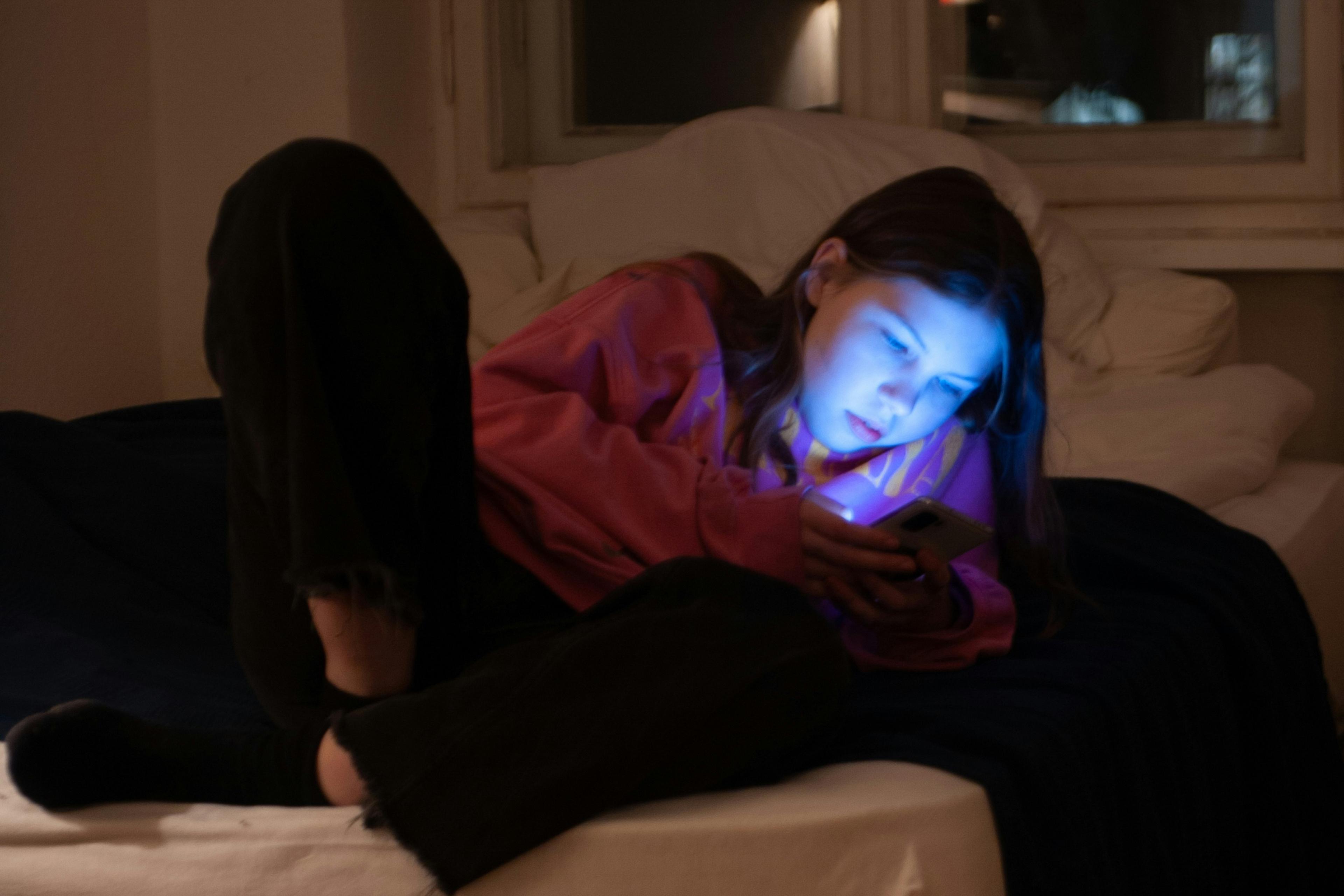 A young girl in a pink hoodie lit by a phone screen, lying on a bed in a dimly lit room at night.