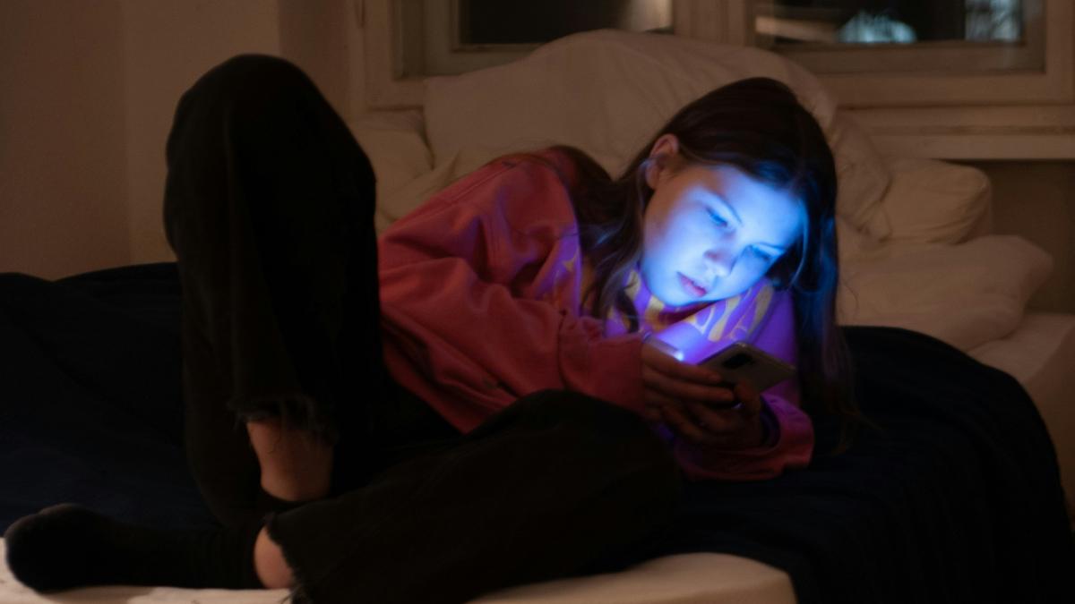 A young girl in a pink hoodie lit by a phone screen, lying on a bed in a dimly lit room at night.