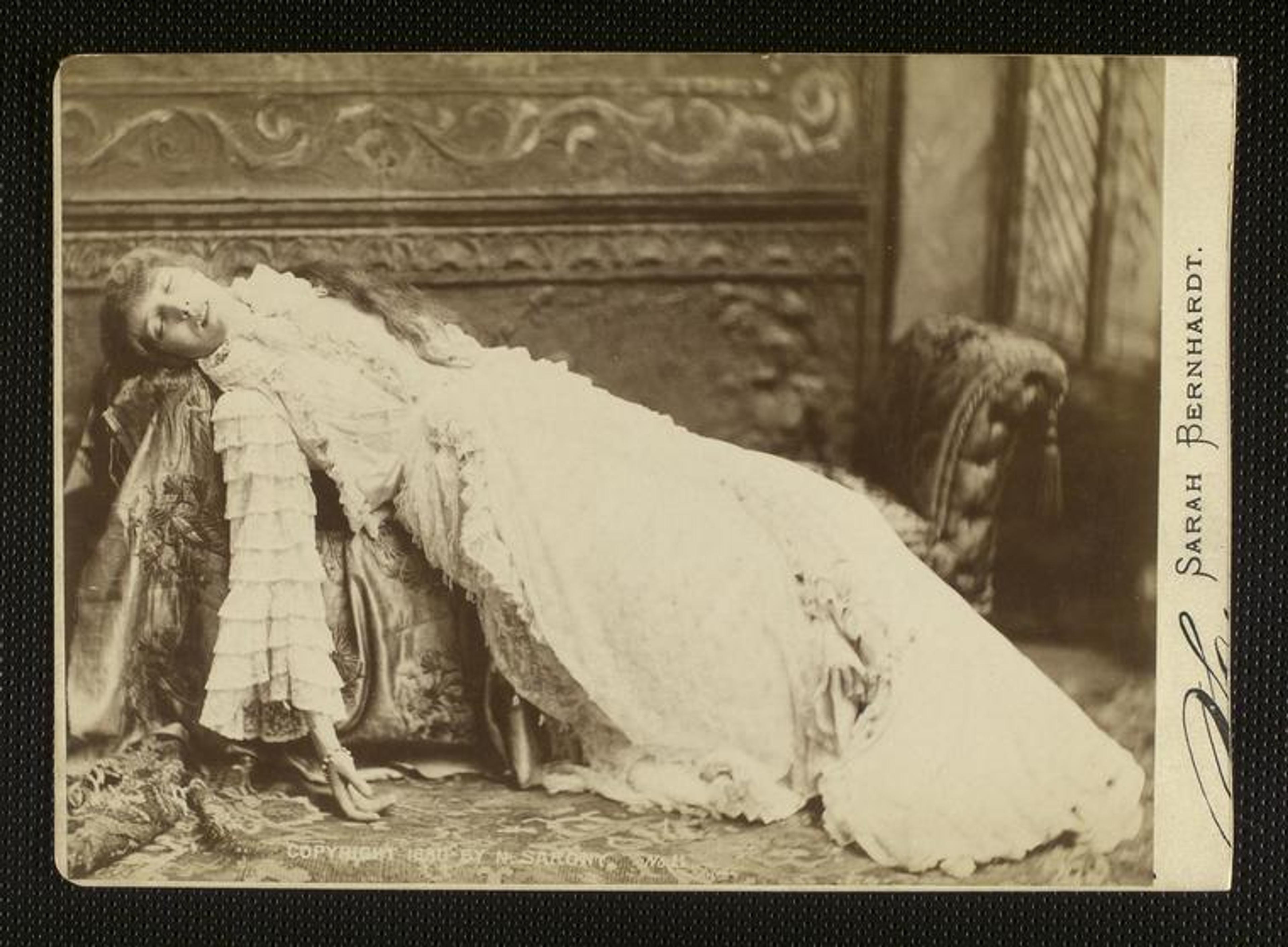 Vintage photograph of a woman in a long, frilly white dress, reclining dramatically on a plush couch with ornate wooden carvings.