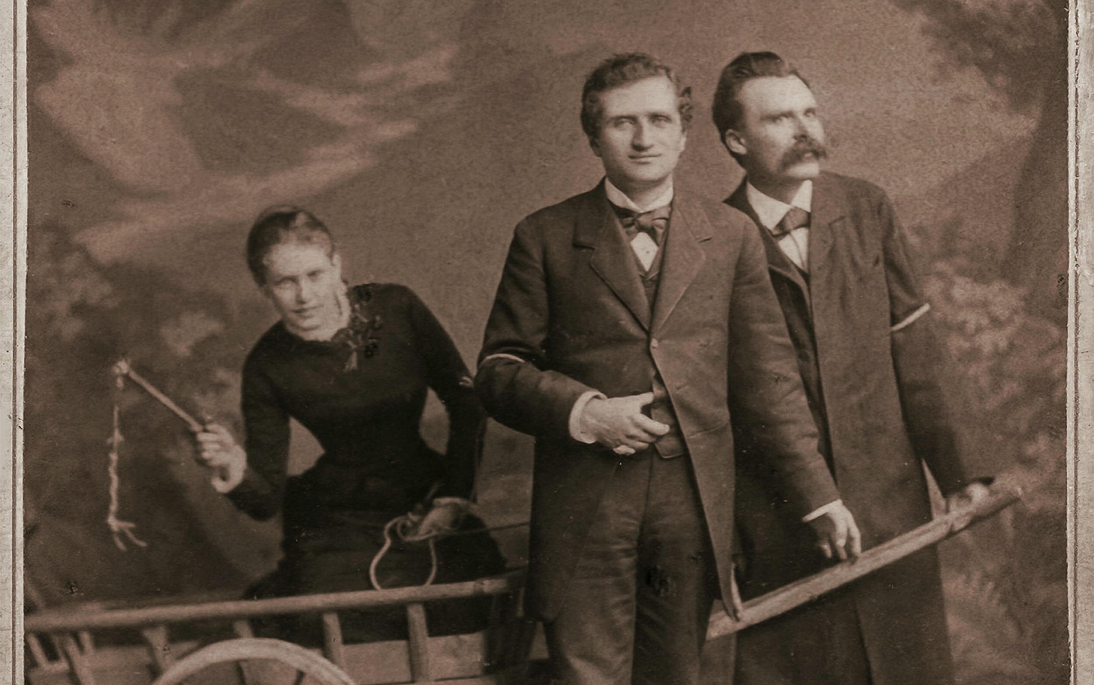 Sepia photograph of two men in suits and a woman in a black dress; the woman holds a whip, leaning out of a wooden cart pushed by the men.