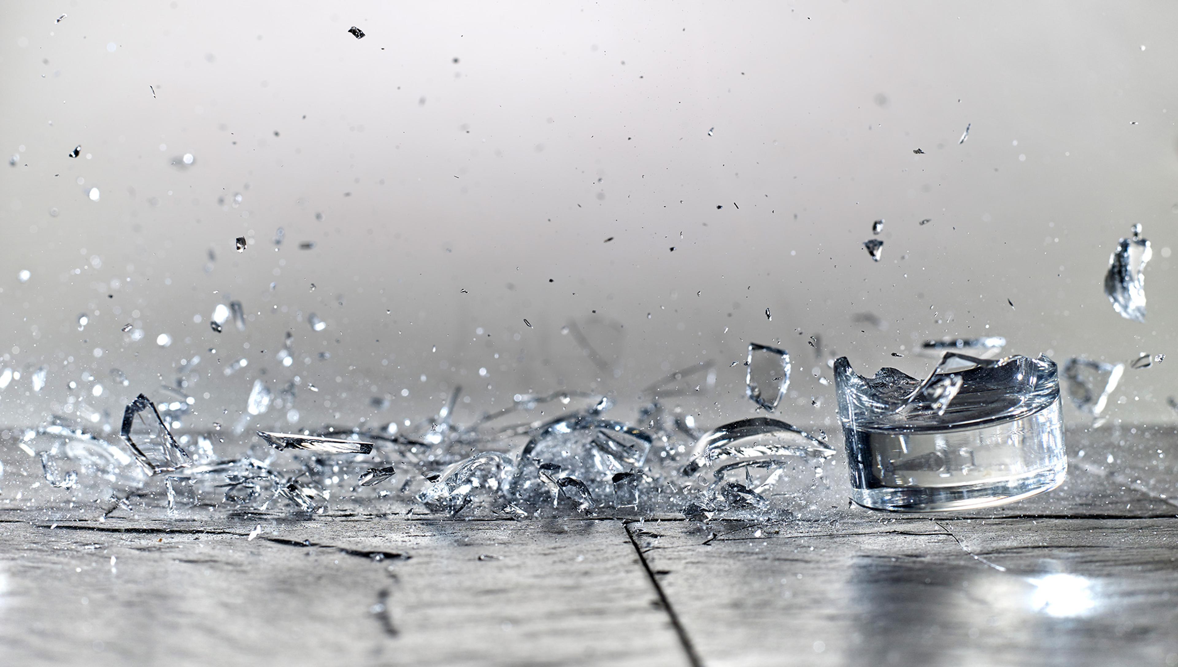 Shattered glass fragments scattered on a grey surface with a blurred background. Some pieces are mid-air, capturing the moment of a glass object crashing and breaking. The image portrays motion and impact.