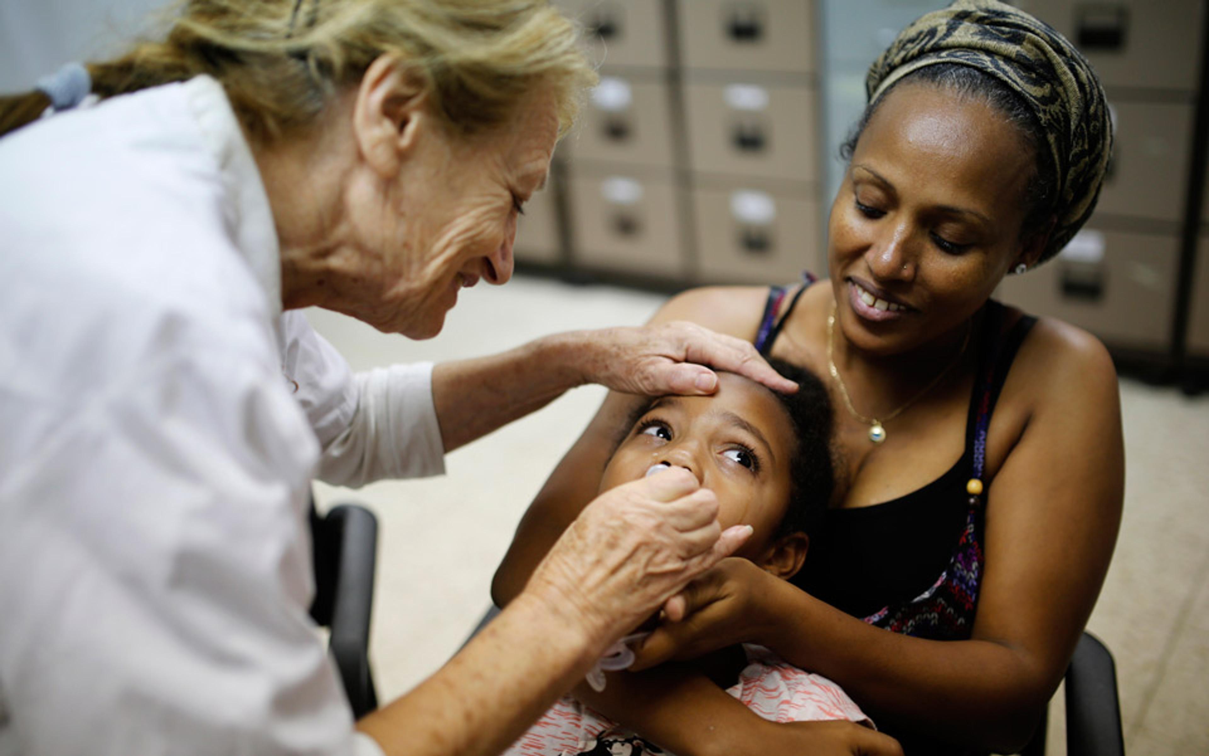 A healthcare worker examining a child, with the child being held by an adult woman in a clinical setting.