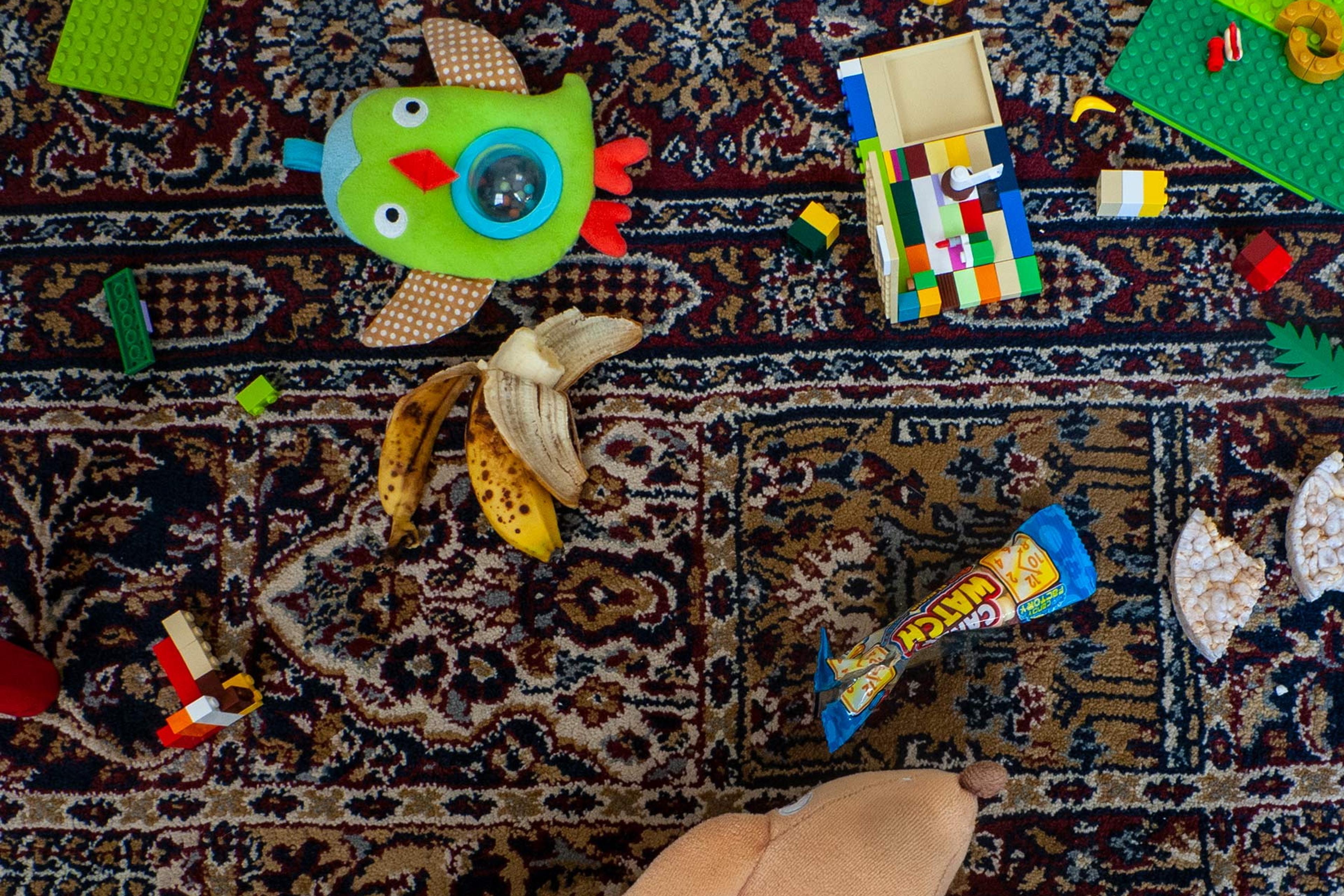Photo of a patterned rug scattered with toys, Lego bricks, banana peel and snacks.