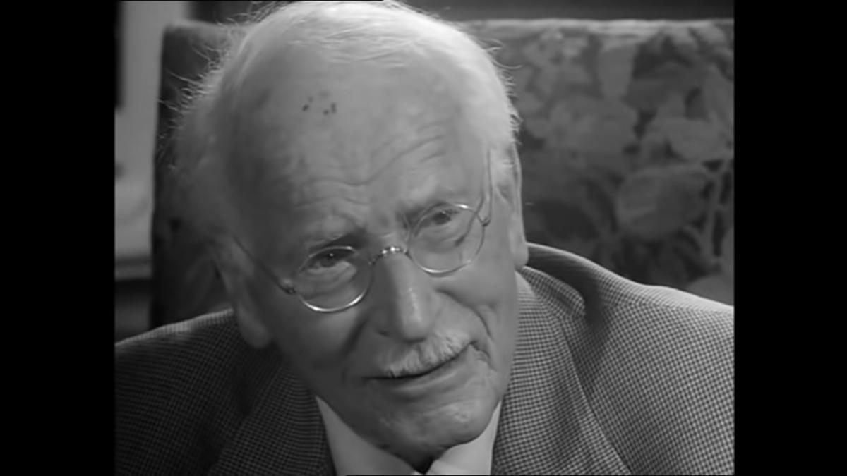 Black and white photo of an elderly man with glasses and a moustache wearing a suit, sitting in a patterned chair.