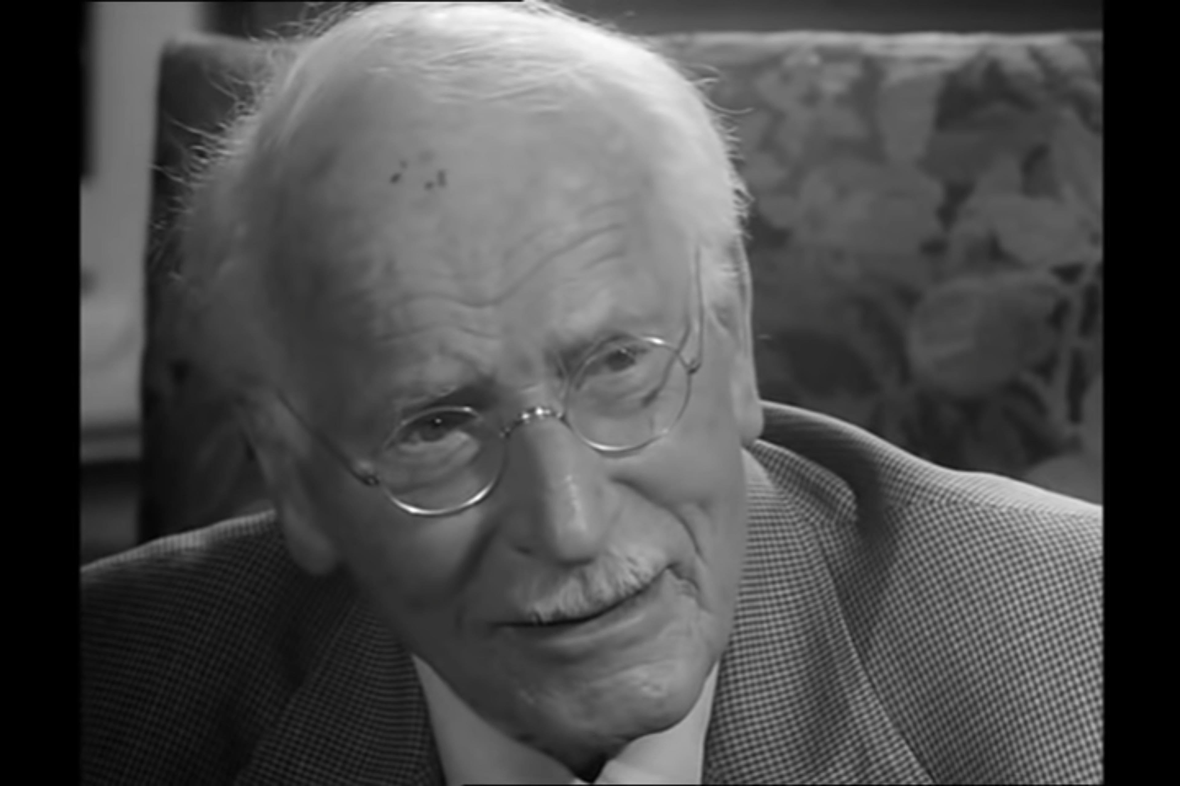 Black and white photo of an elderly man with glasses and a moustache wearing a suit, sitting in a patterned chair.