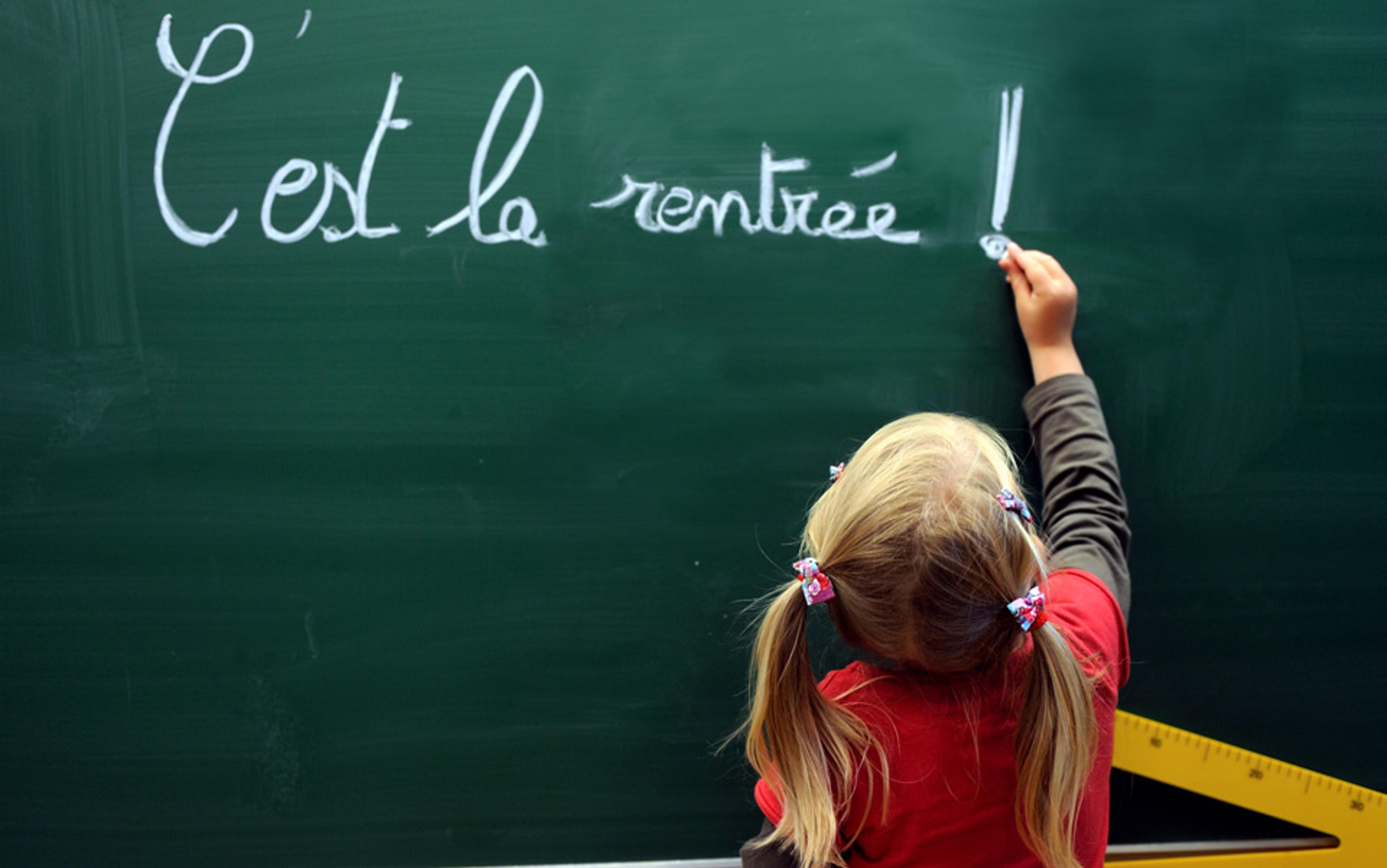 A little girl with blonde hair writing “C’est la rentrée !” in chalk on a green chalkboard, signifying the start of the school year.